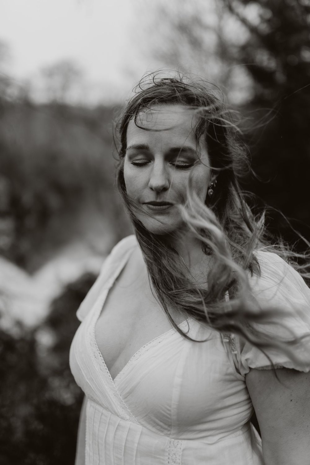 Black and white portrait of a woman in a white dress with windswept hair and closed eyes outdoors.