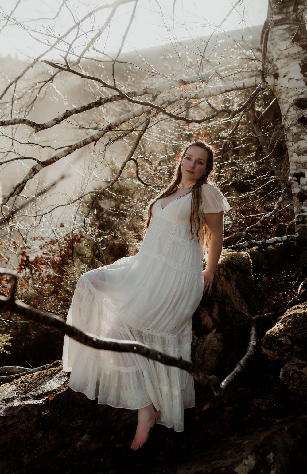 Young woman in white flowing dress poses barefoot on rocks among bare trees in a misty forest setting.