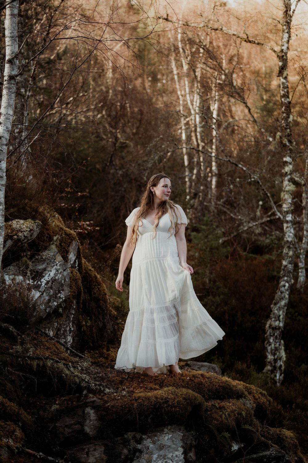 Woman in white flowing dress standing among mossy rocks and birch trees in an enchanted forest setting.