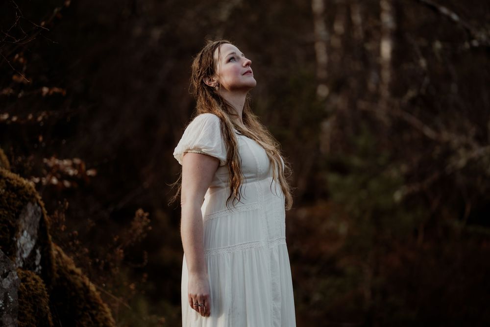 Woman in white dress gazing upward in a moody forest setting with dark trees and rocks.