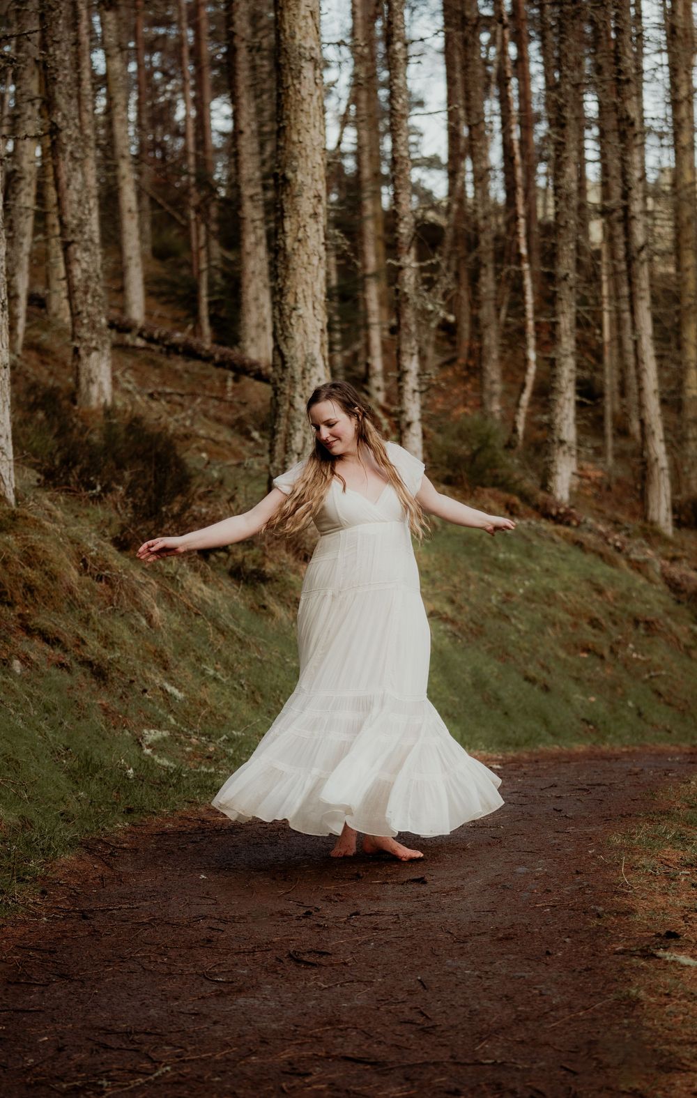 A woman in a flowing white dress dances barefoot on a forest path surrounded by tall trees.