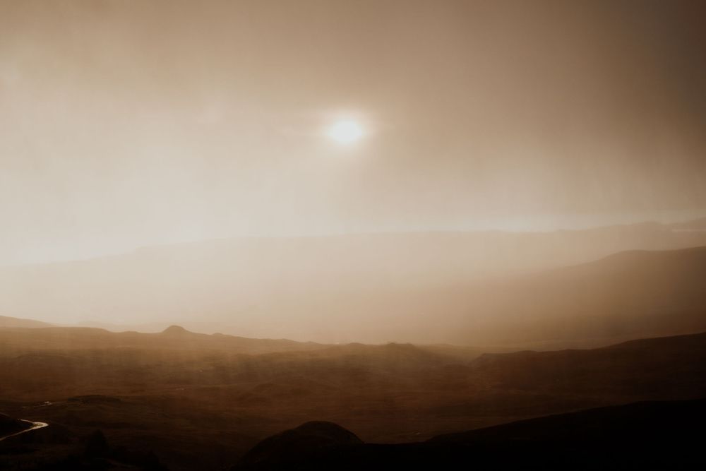 Hazy Mars-like desert landscape with a glowing sun through dusty orange fog over silhouetted mountain ridges.