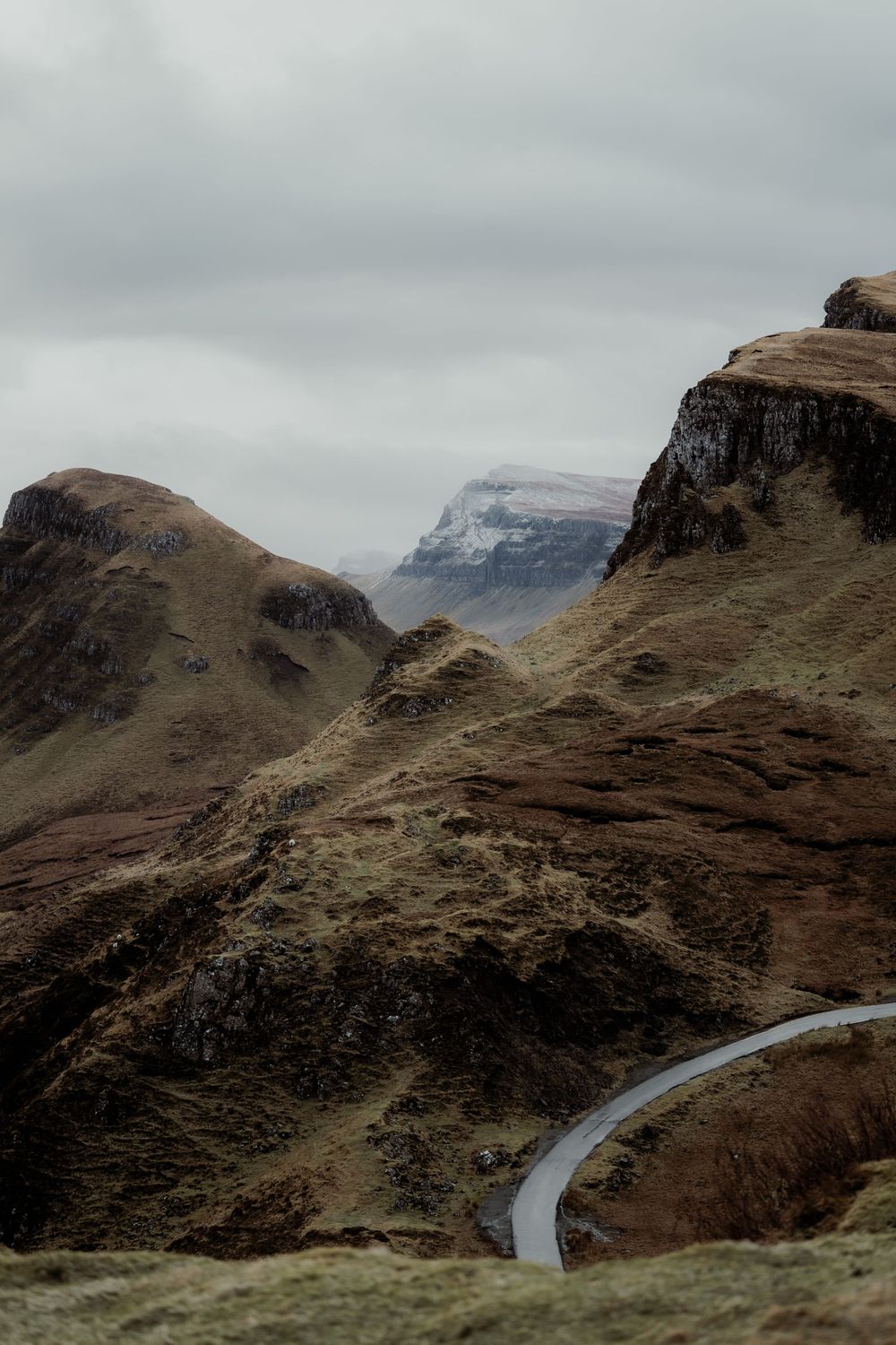A winding road curves through dramatic rocky highland mountains under a cloudy grey sky.