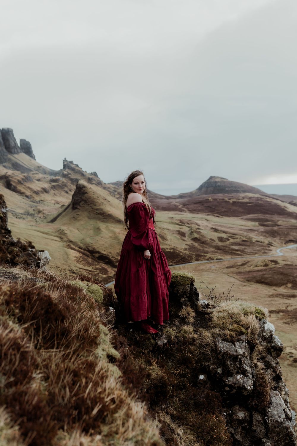 A woman in a red off-shoulder gown stands on rocky highland terrain with misty mountains behind.
