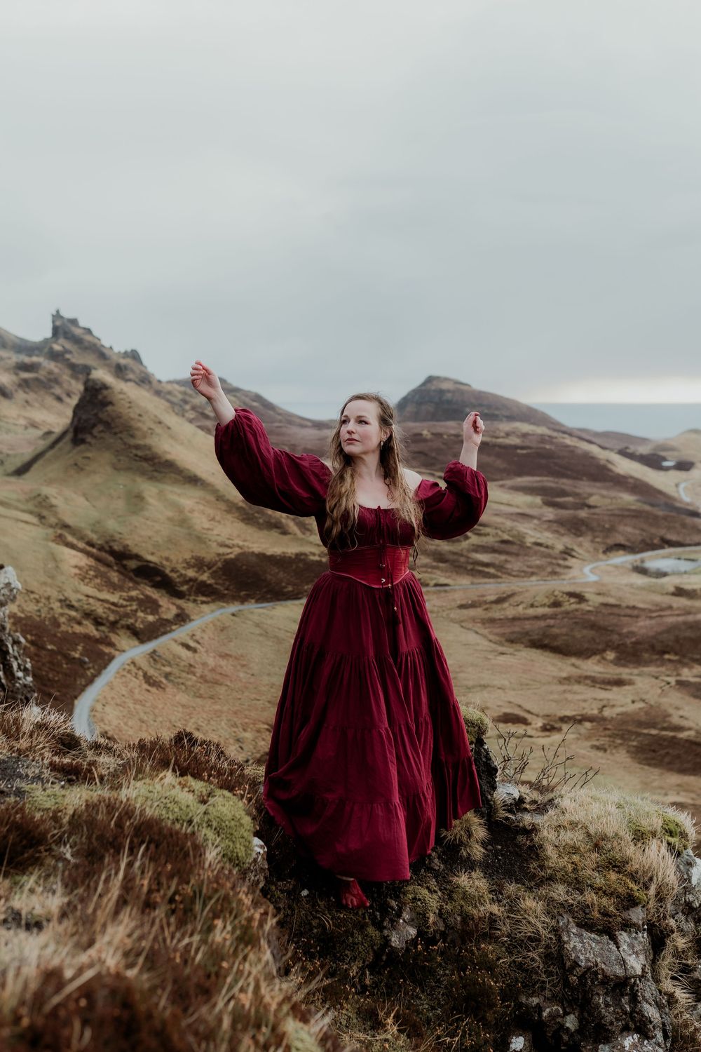Woman in red medieval gown poses dramatically on Scottish Highland hills with arms raised.