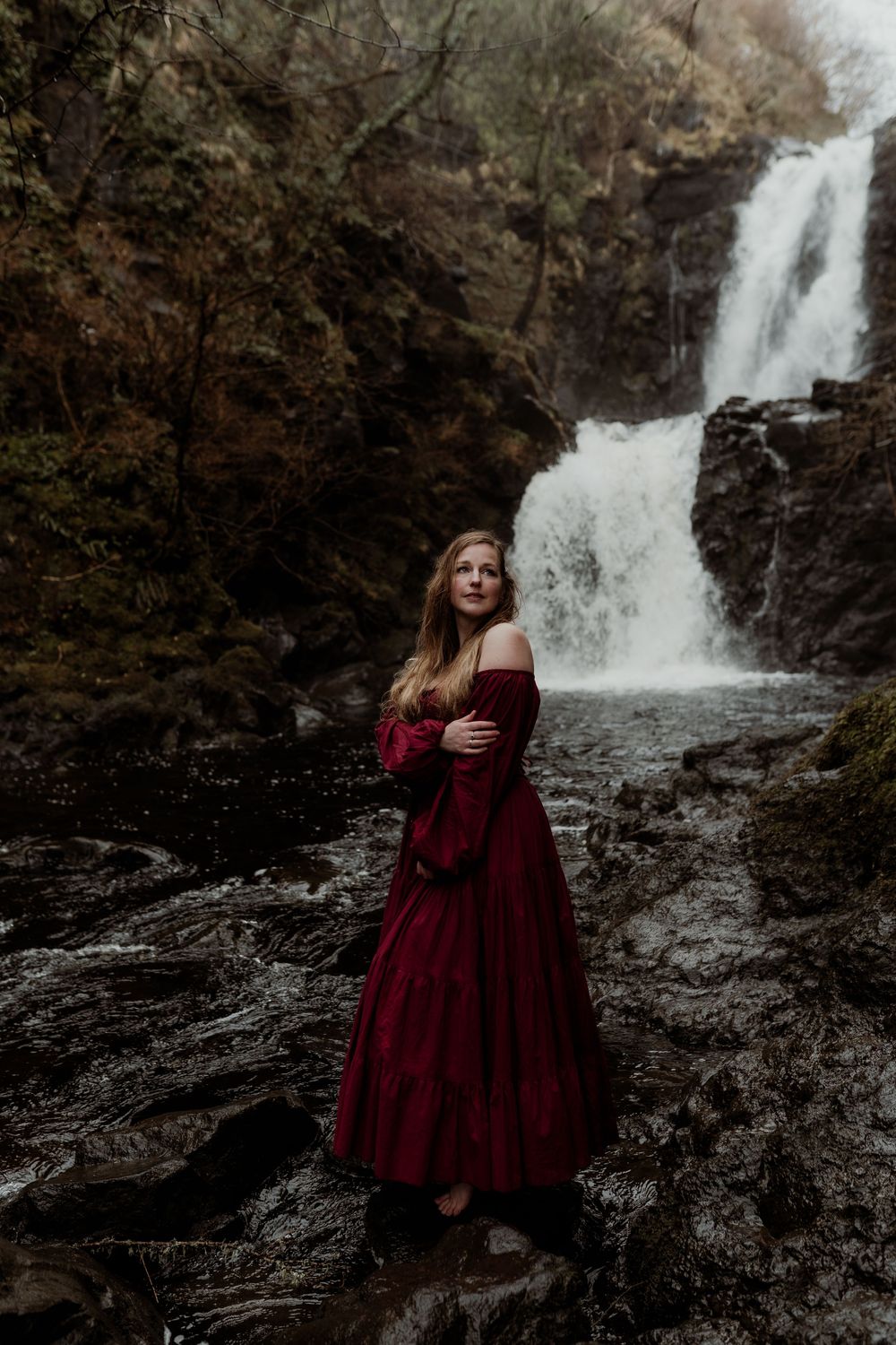 Woman in red off-shoulder gown stands before a cascading waterfall in a forest.