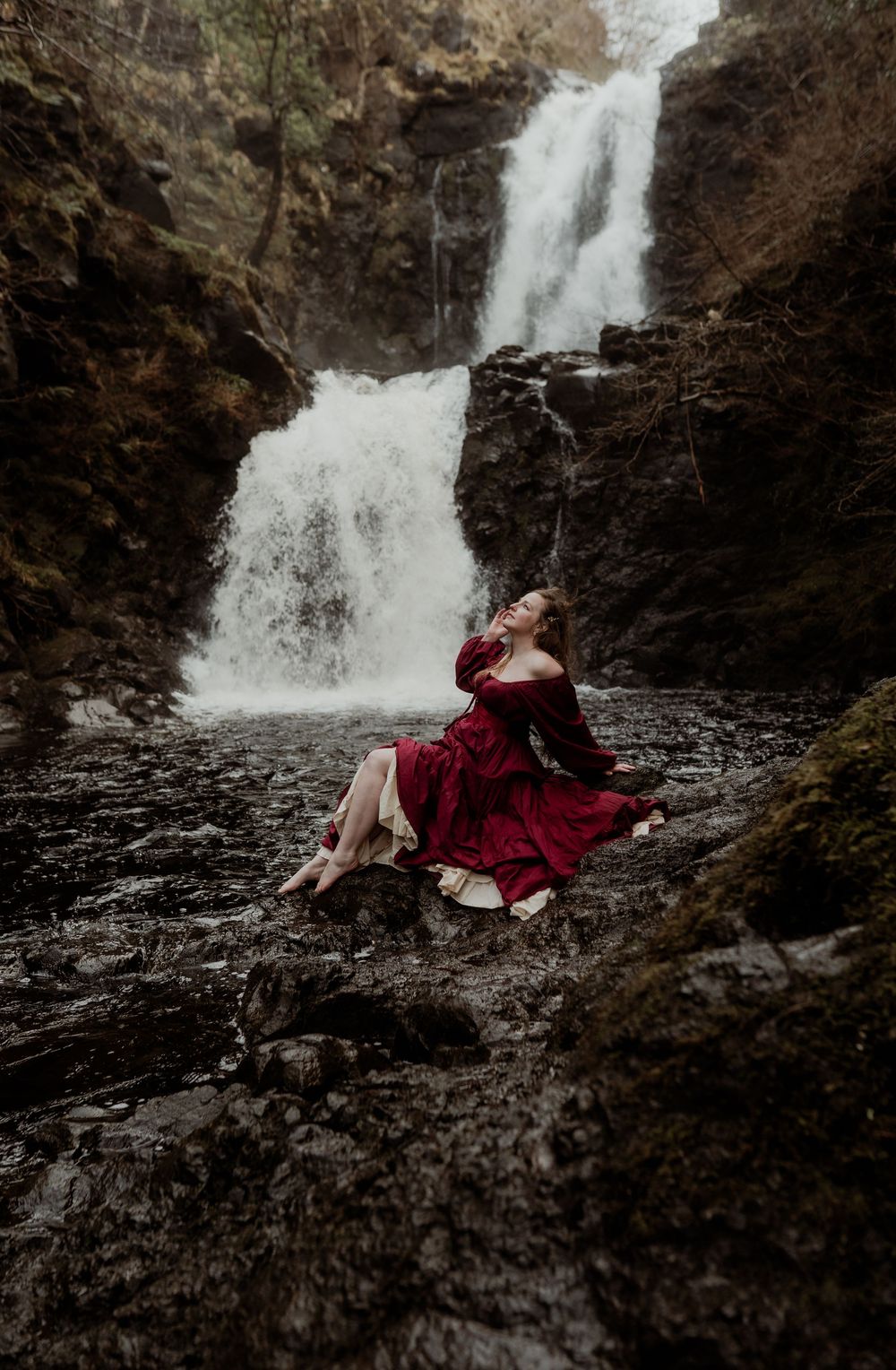 Woman in red dress sitting on rocks before a cascading waterfall in a moody forest setting.