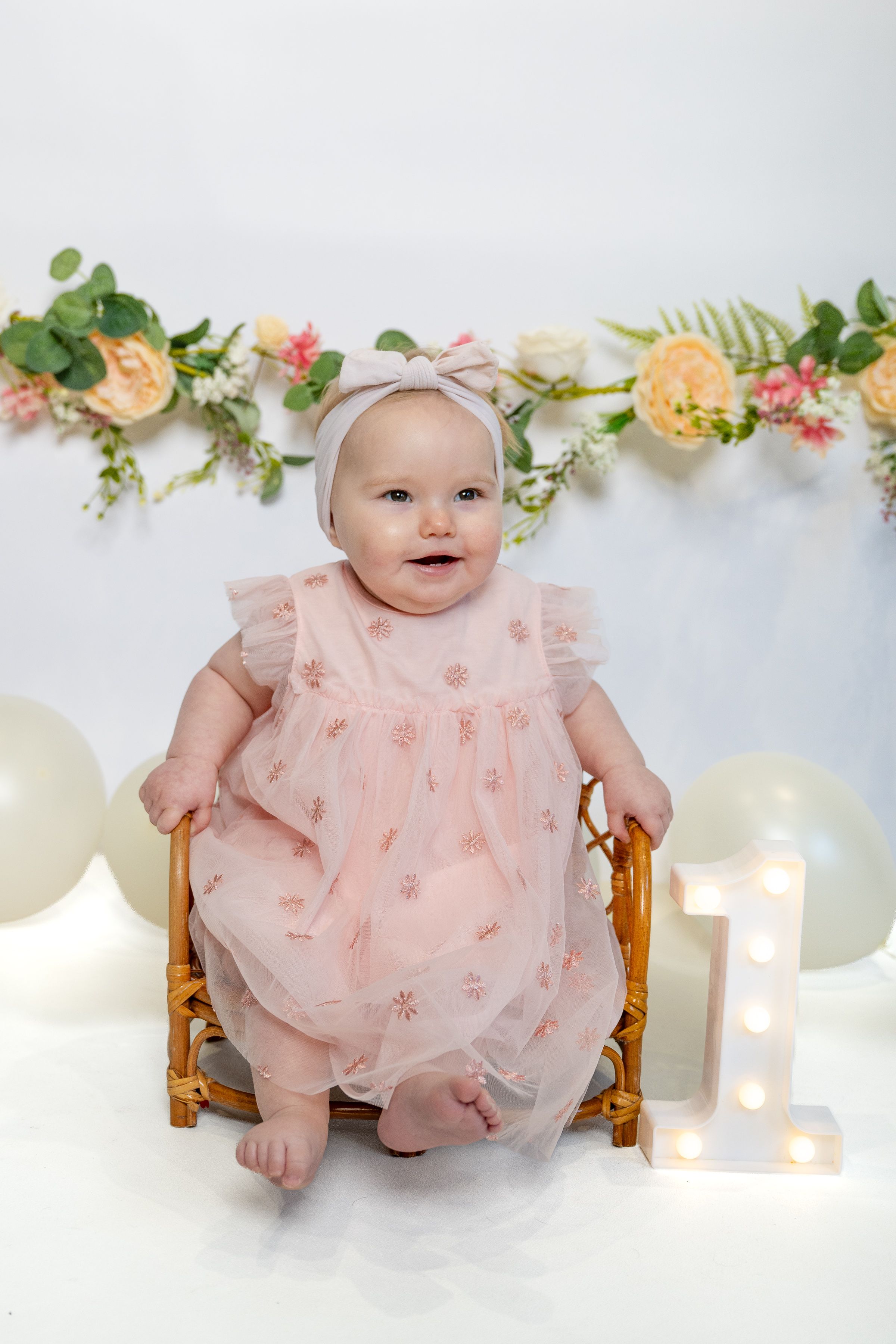 Baby girl in pink tulle dress sits in rattan chair beside illuminated number 1, celebrating her first birthday.