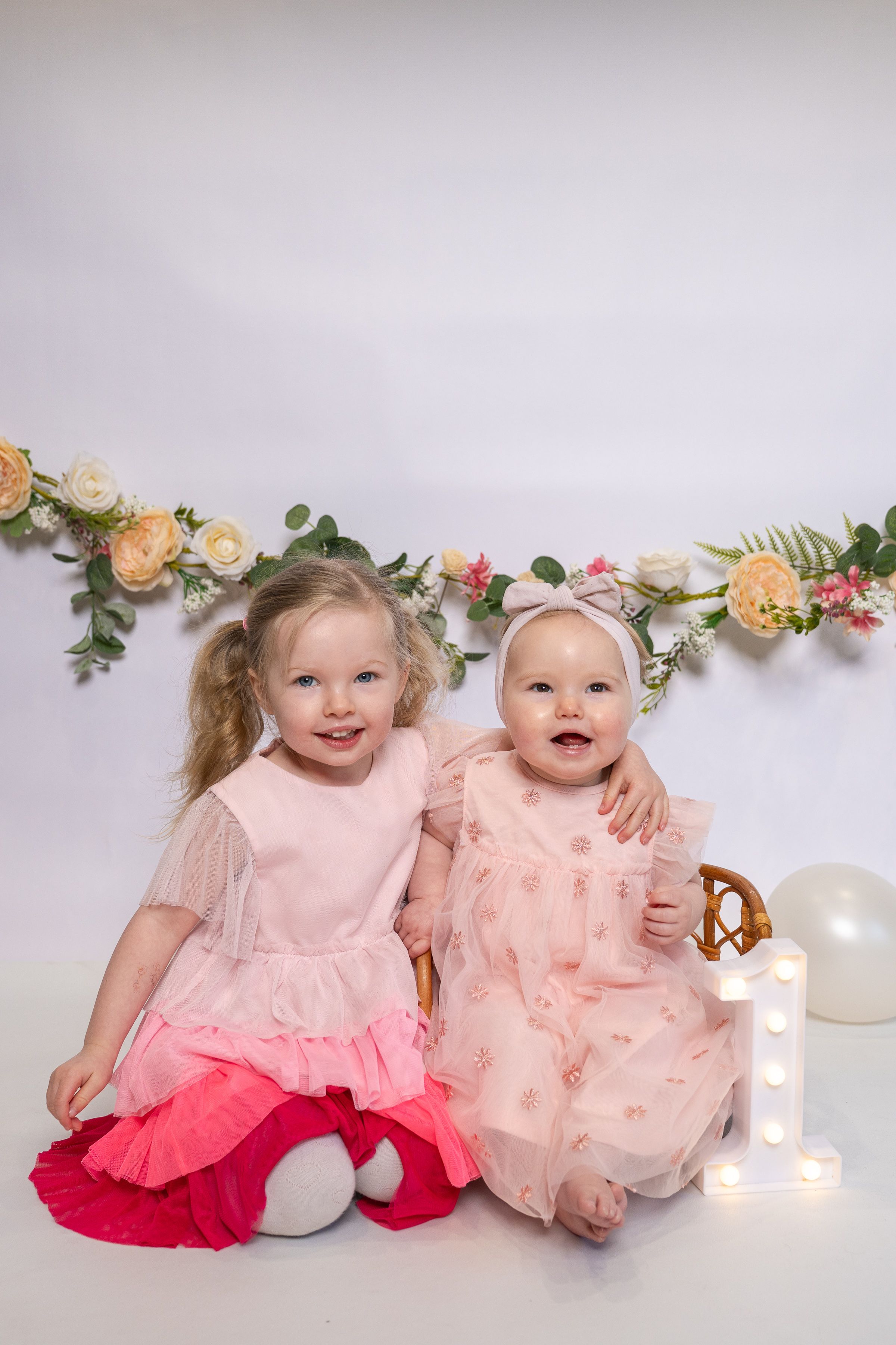 Two young girls in pink dresses pose together with floral garland backdrop and illuminated number one decoration.