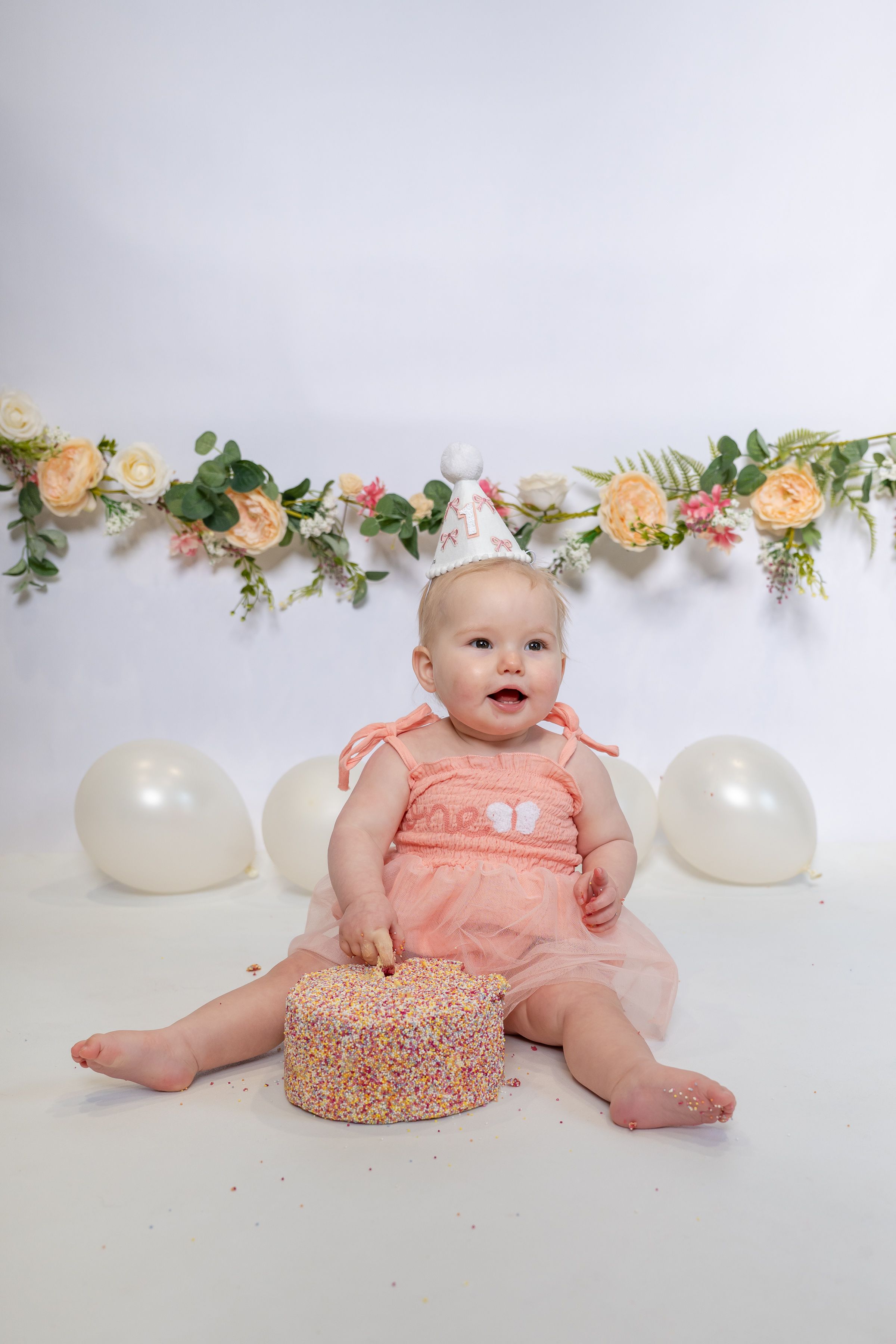 Baby girl in pink dress and party hat sits with smash cake, balloons and floral garland for first birthday photo.