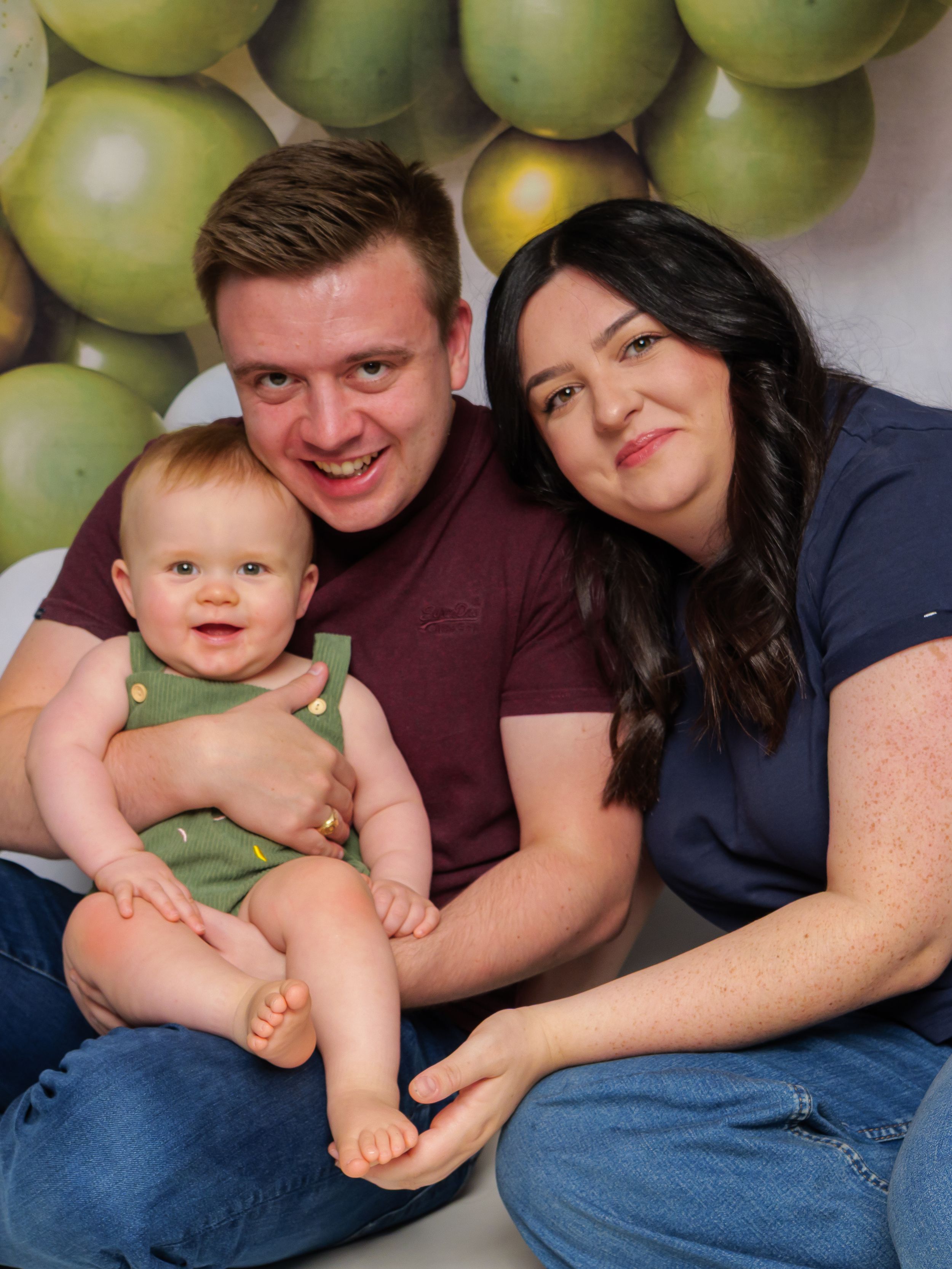 Happy family portrait with parents and smiling baby in green overalls, posed in front of green balloon backdrop.