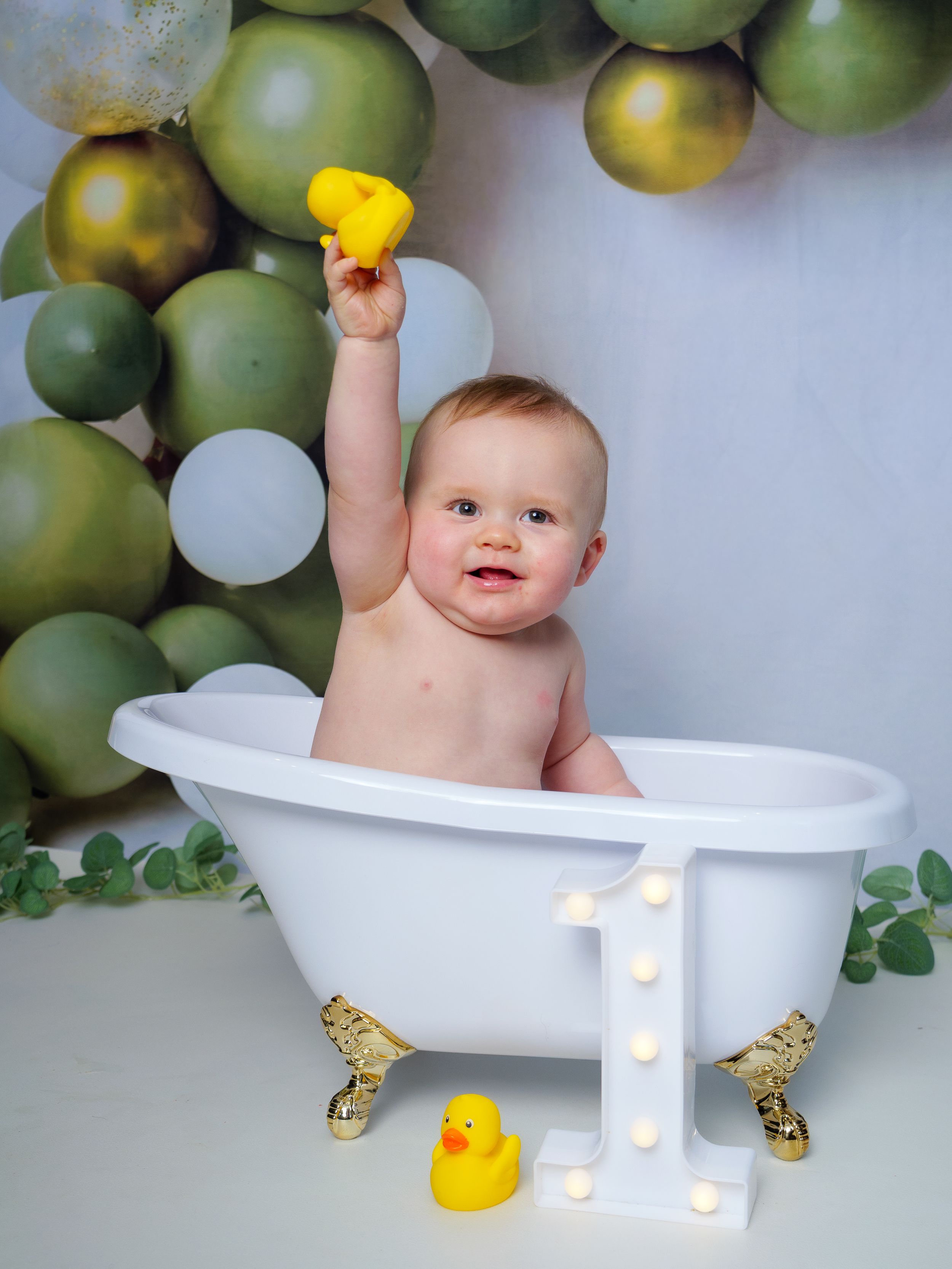 Baby in a white clawfoot tub holds a yellow rubber duck, surrounded by green balloons and a lit number one sign.