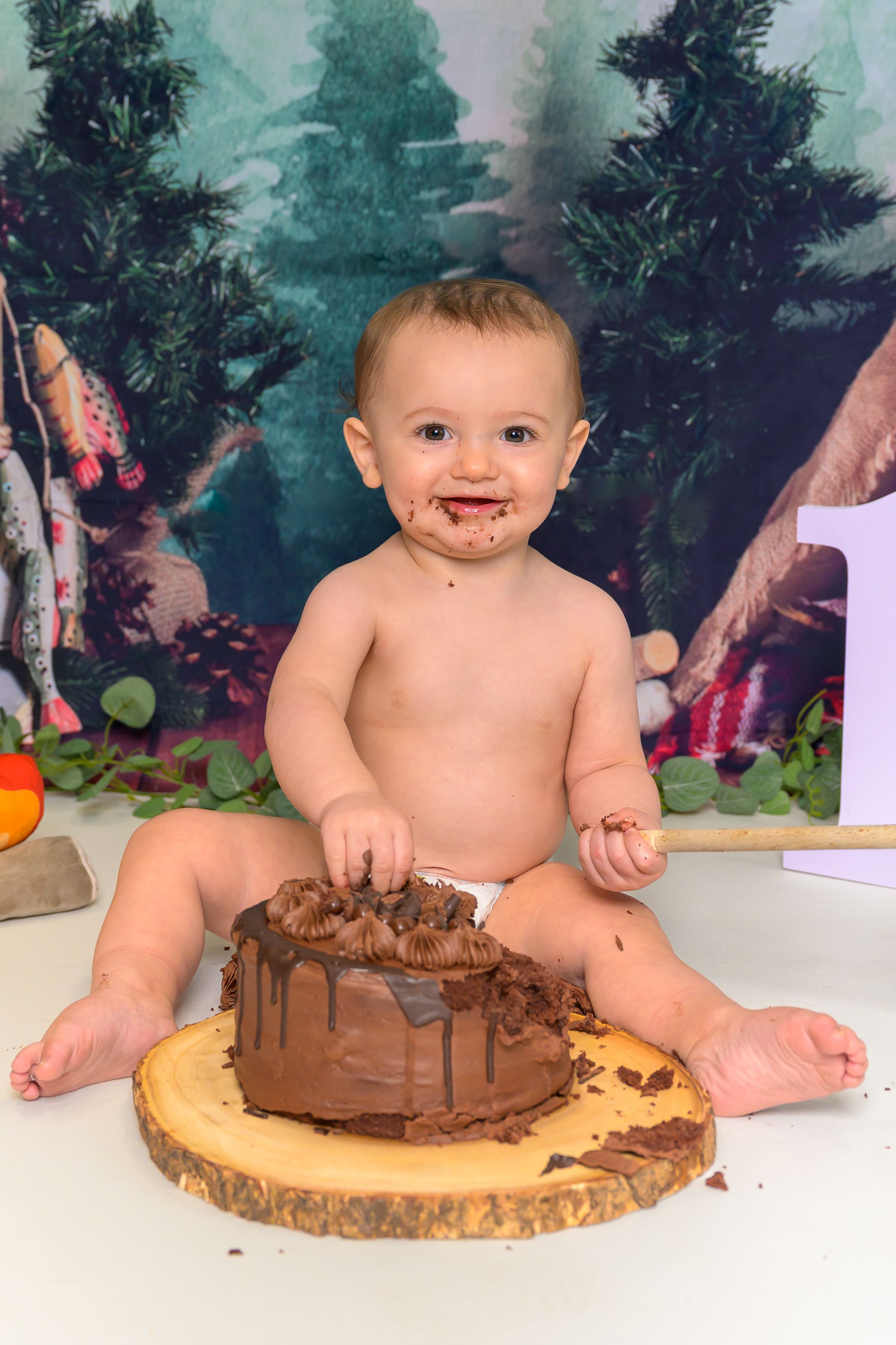 Smiling baby covered in chocolate frosting during a first birthday cake smash with a forest backdrop.