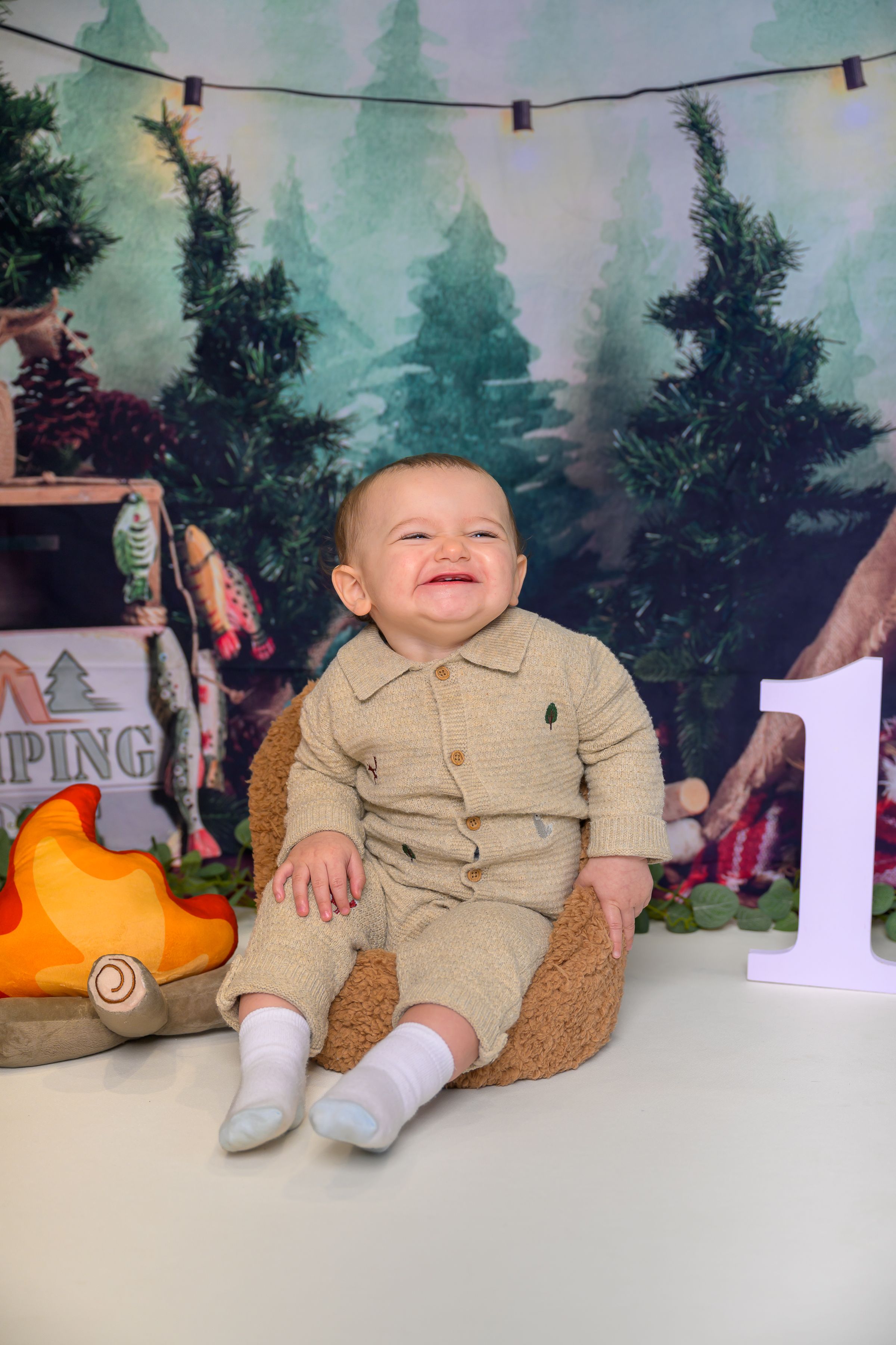 Smiling baby in beige outfit sits in mini chair with camping-themed backdrop and number 1 for first birthday photo.