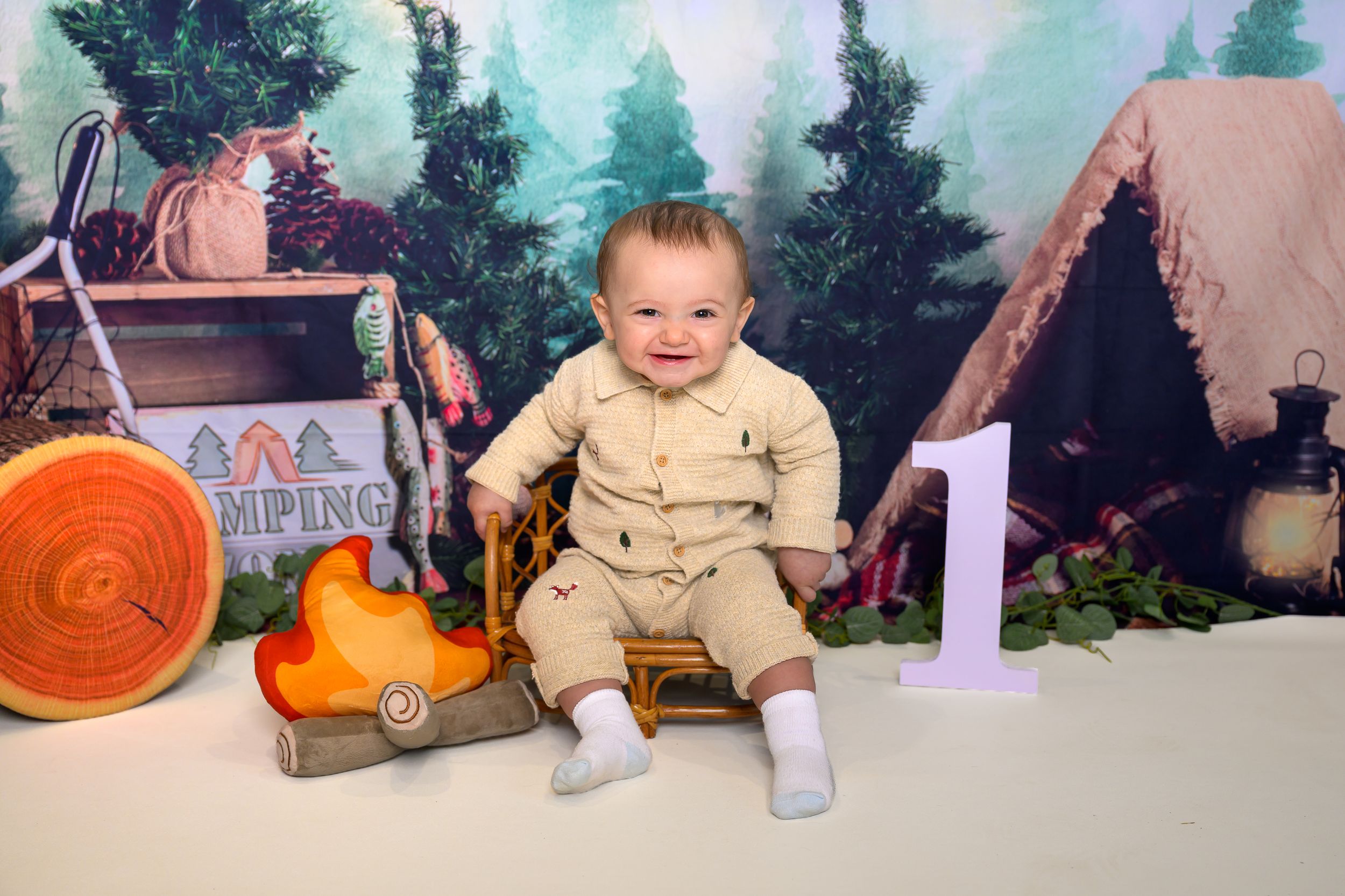 Baby boy in beige outfit sits smiling at a camping-themed first birthday photo shoot with trees and tent backdrop.
