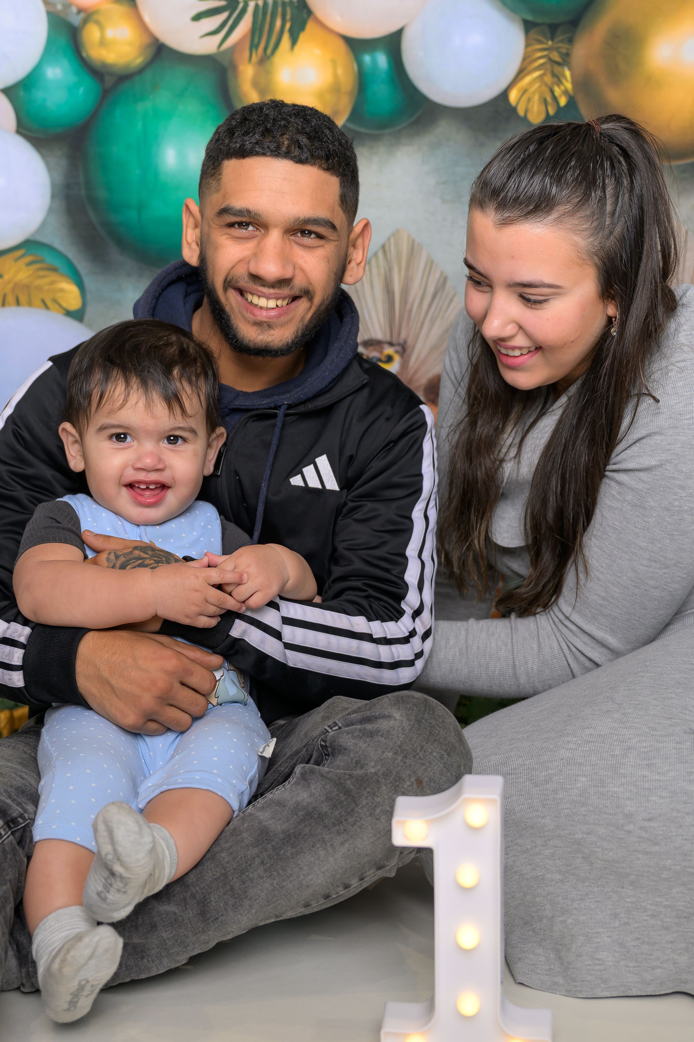 A young family celebrates their baby's first birthday with a number one light-up sign and colorful balloon backdrop.