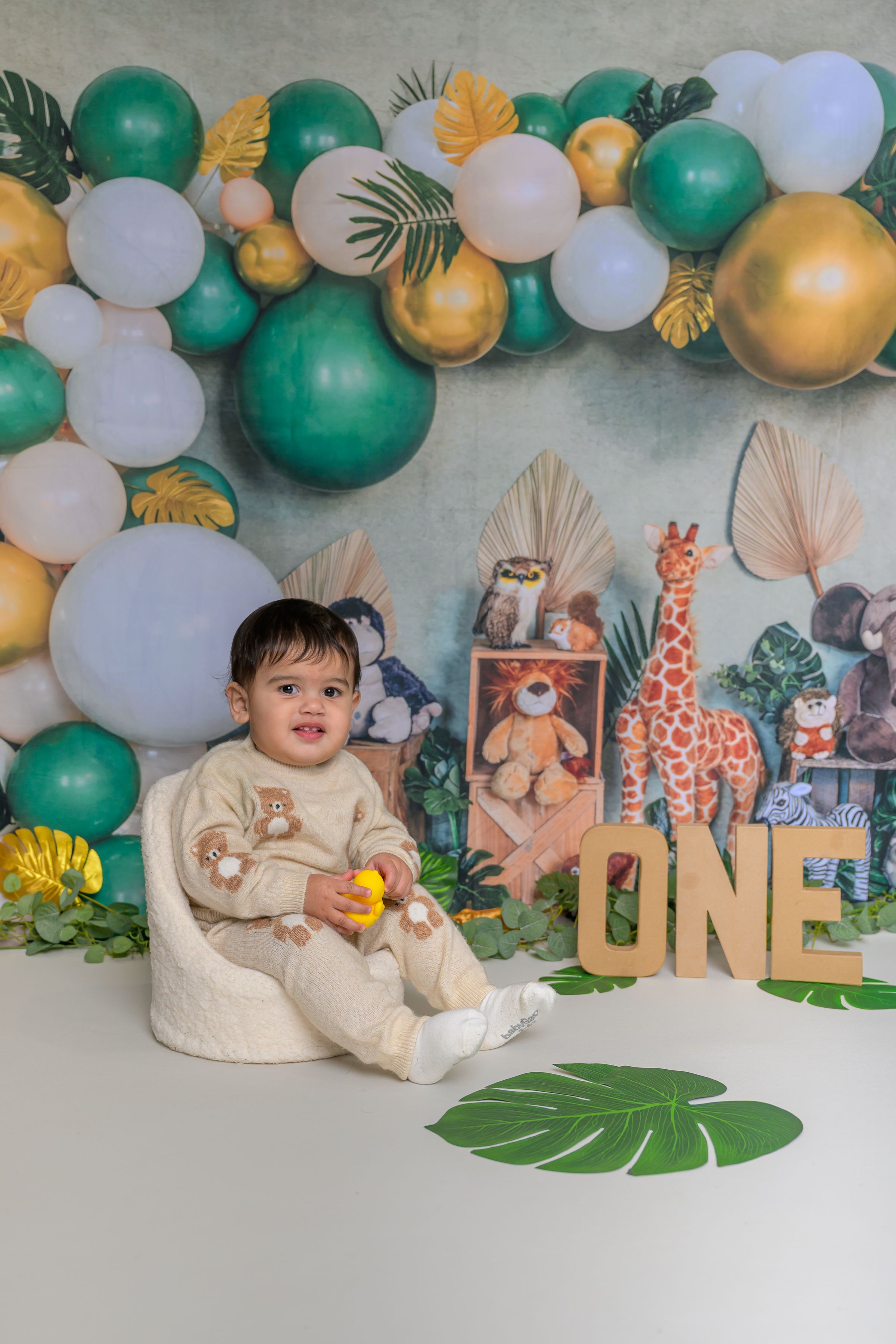 Baby's first birthday photo with safari theme, green and gold balloons, stuffed animals, and ONE sign.