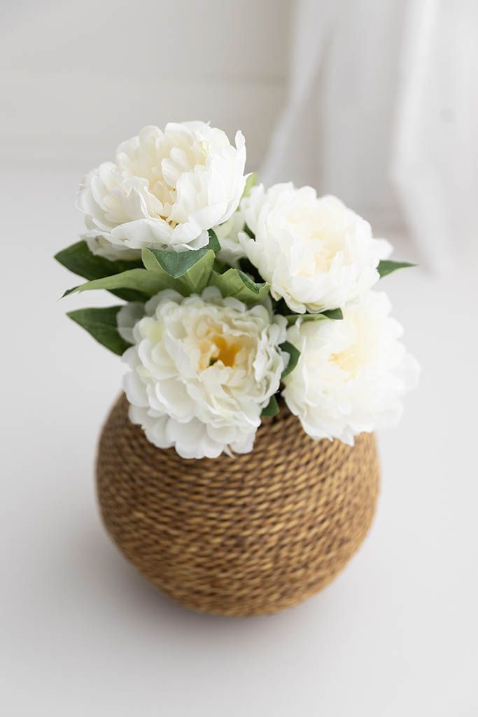 White peony flowers arranged in a round rope-wrapped vase on a white surface.