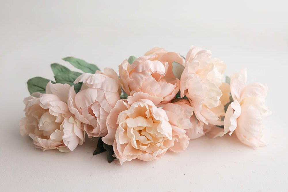 Soft blush pink artificial peony flowers with green leaves arranged on a white background.