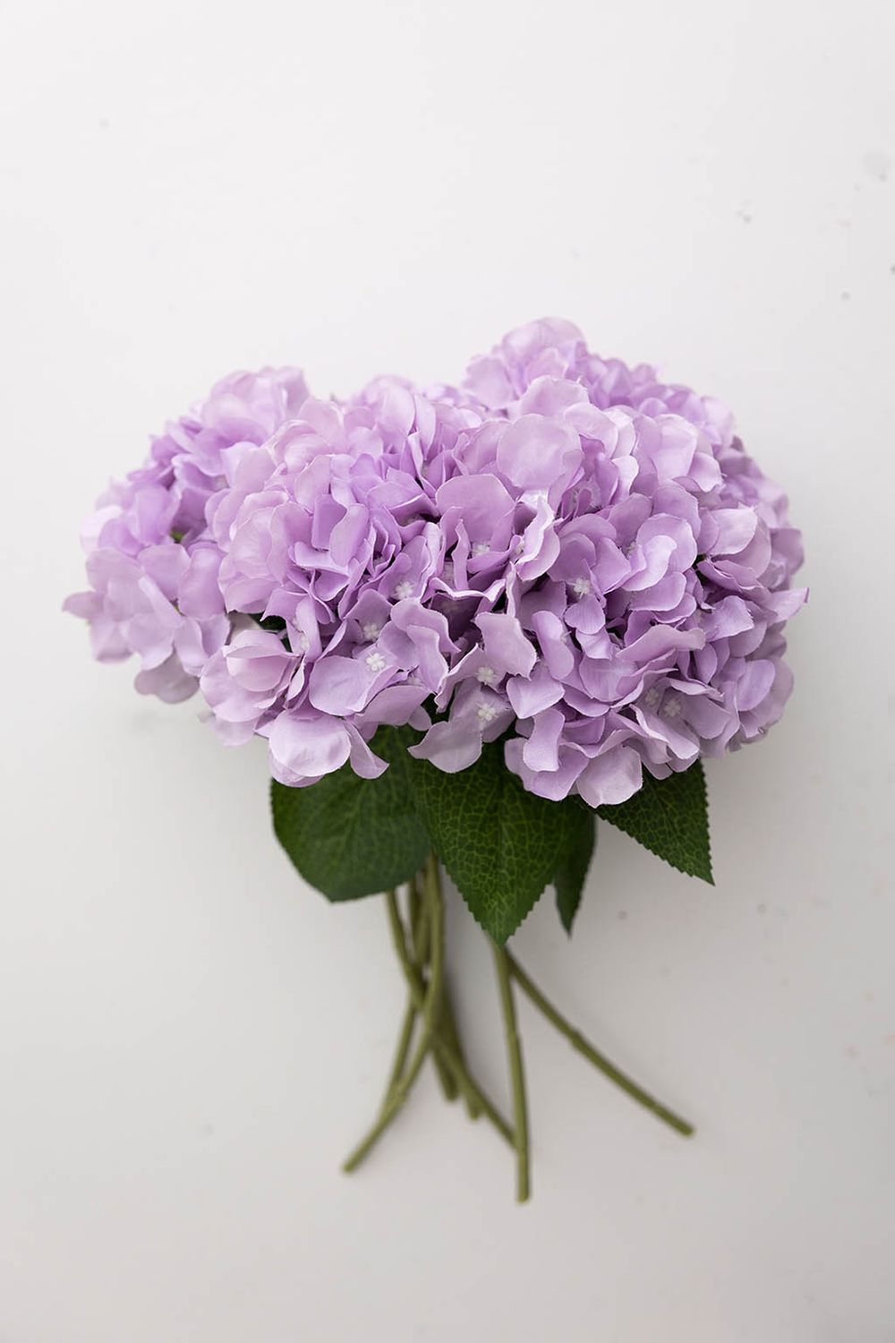 Light purple artificial hydrangea bouquet with green stems on white background.