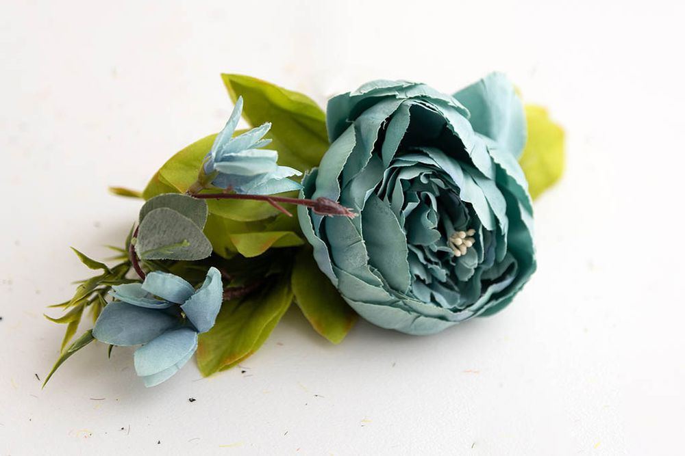 Teal blue artificial peony flower with green leaves on a clean white background.