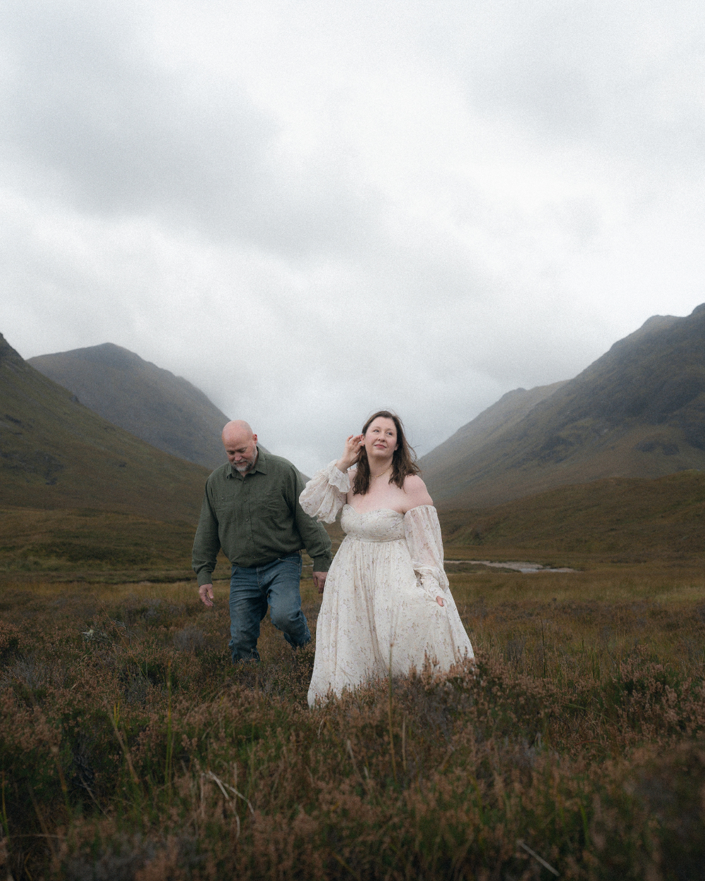 A couple walks hand-in-hand through a misty Scottish Highland valley, woman in white lace dress, man in green jacket.