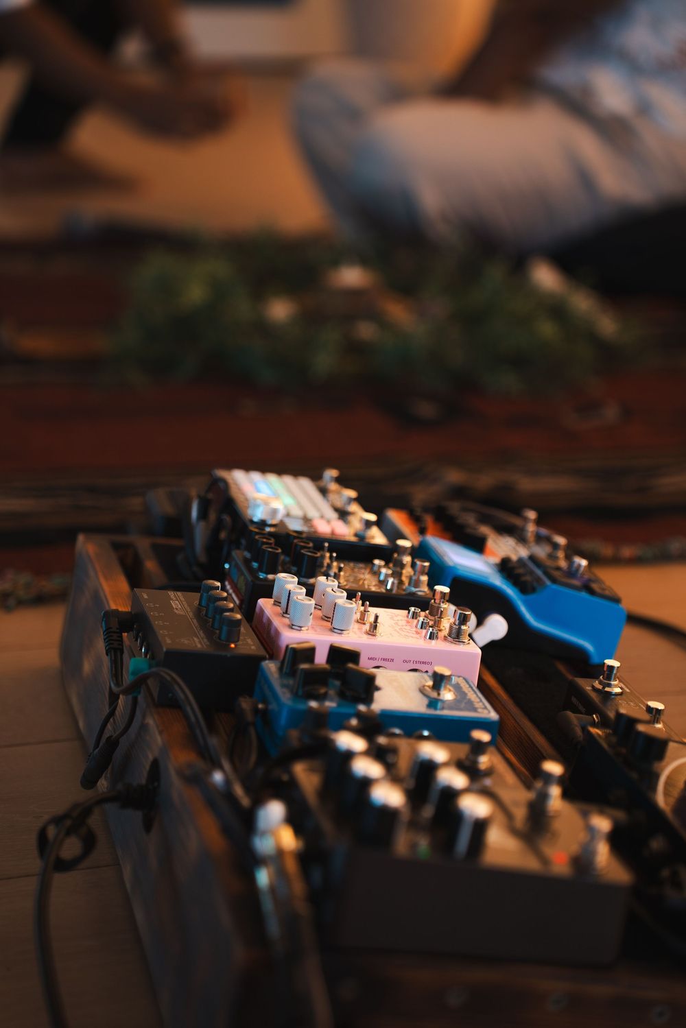Guitar effect pedals arranged on a wooden floor in warm light, with a person sitting in the background. Vista, CA