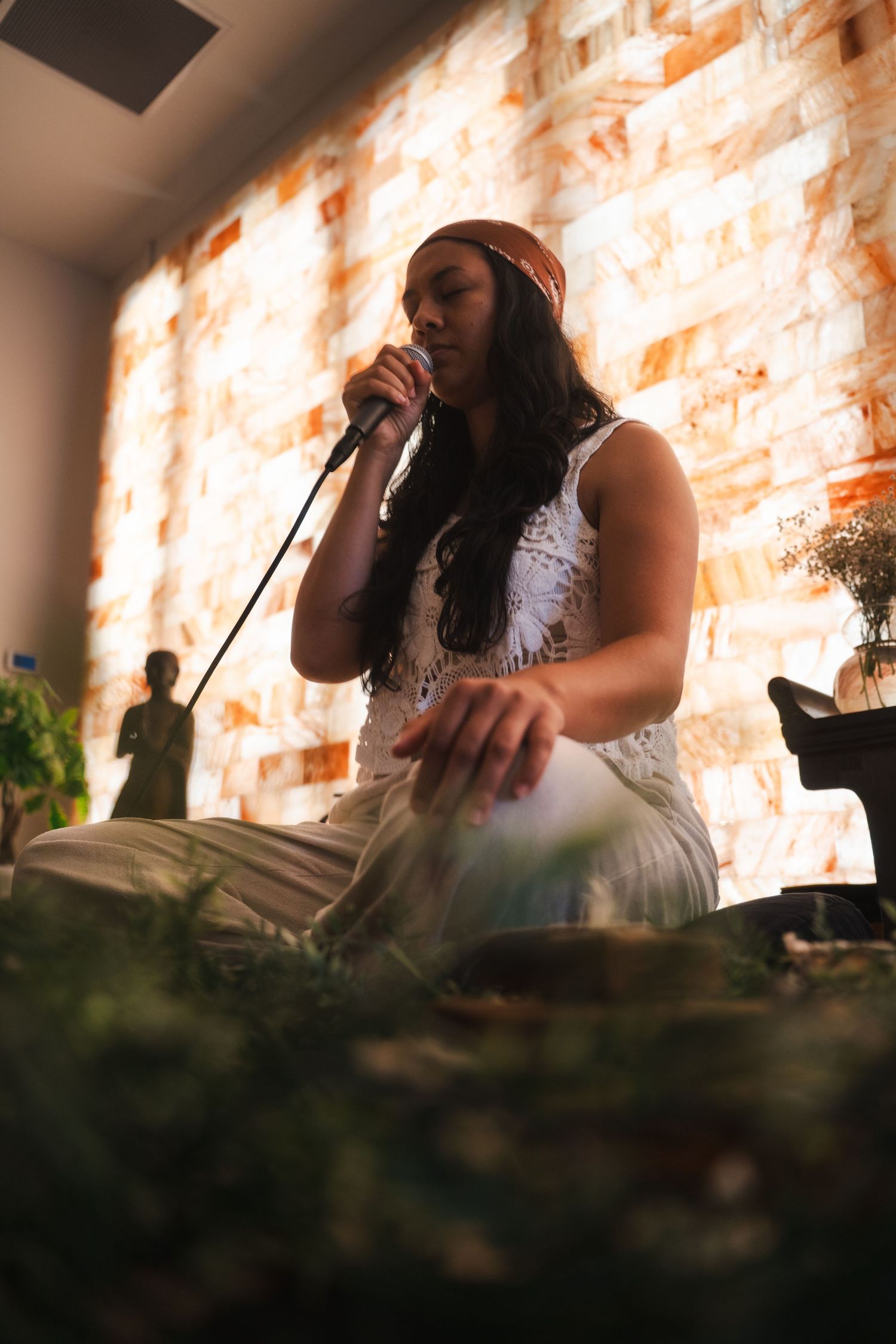 A woman leading meditation while sitting cross-legged surrounded by plants in a warmly lit room. Vista, CA