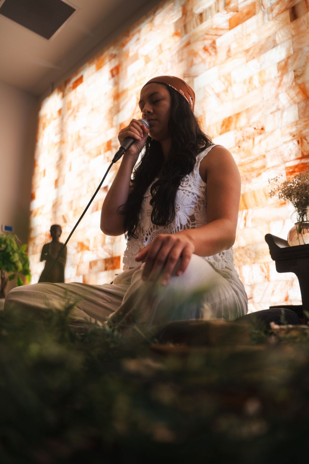A woman leading meditation while sitting cross-legged surrounded by plants in a warmly lit room. Vista, CA