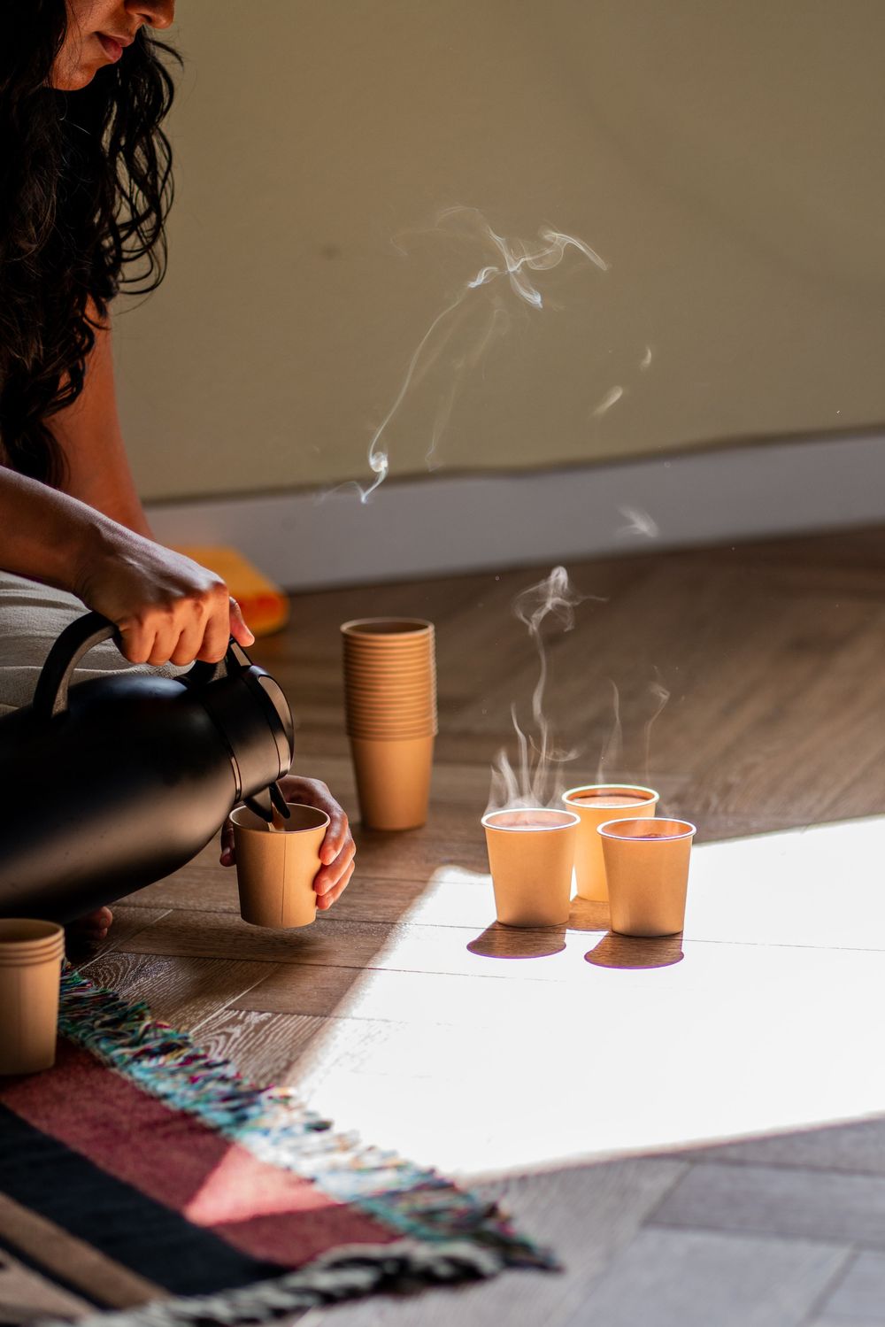 Woman pouring hot coco from black kettle into steaming cups on sunlit wooden floor with colorful rug. Carlsbad, CA
