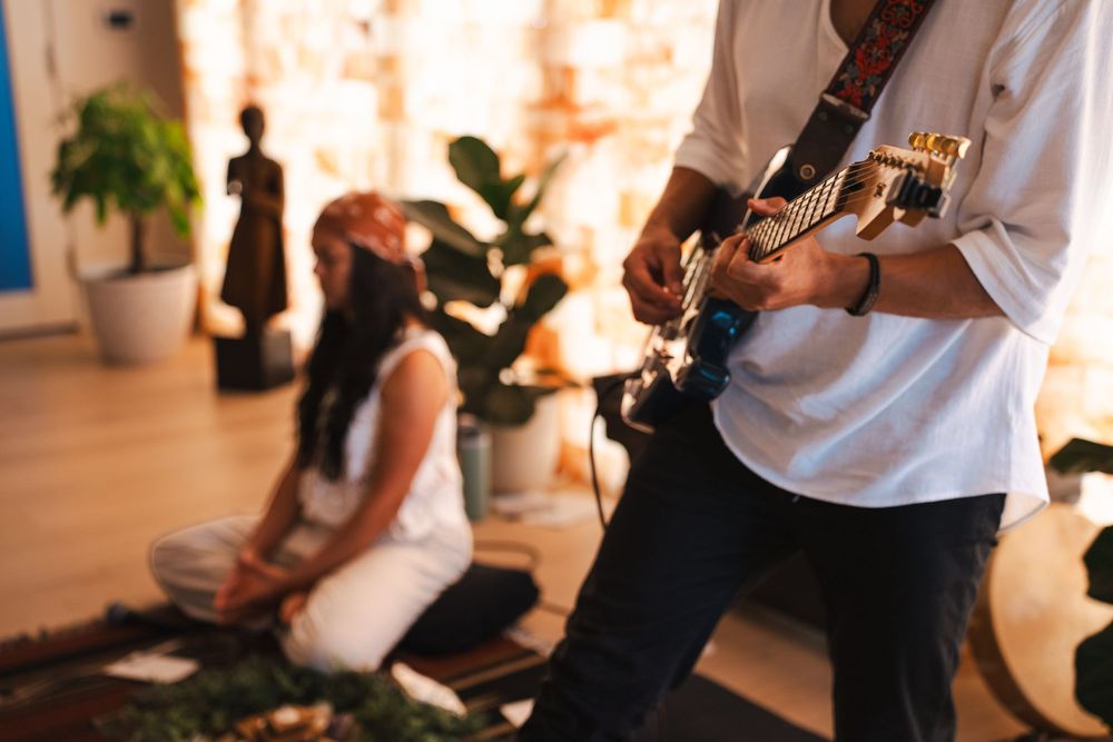 Guitarist in white shirt plays acoustic guitar while a woman sits cross-legged on floor in warmly lit room during guided meditation. Vista