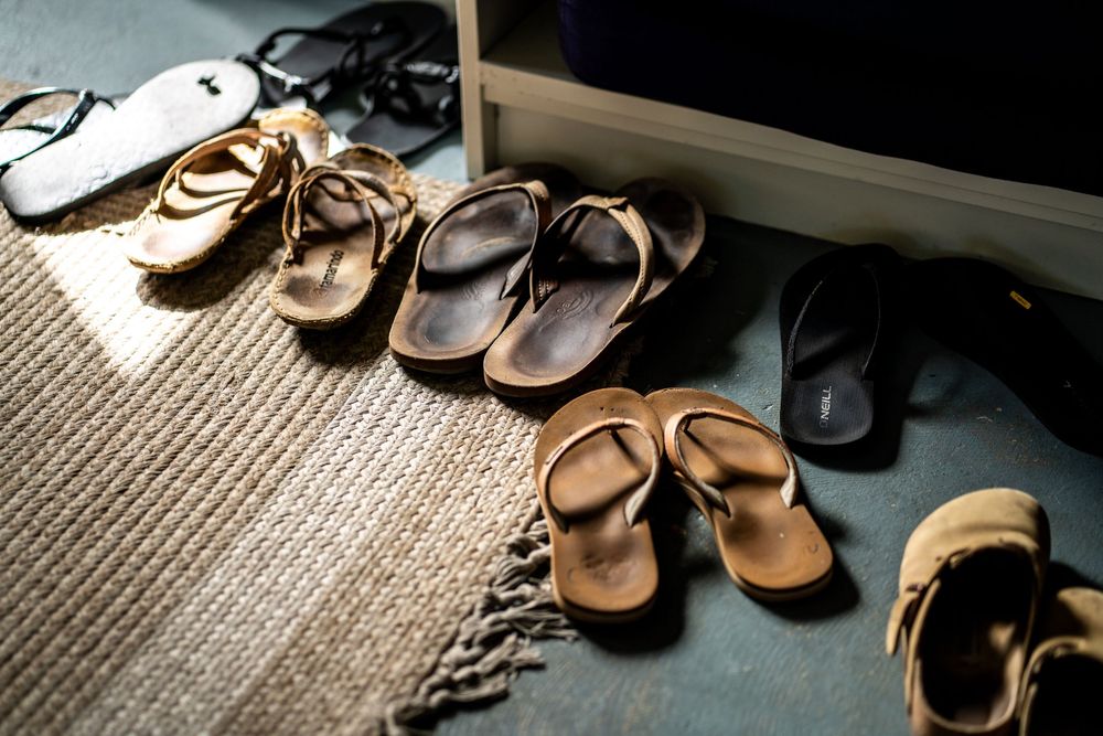 Multiple pairs of worn flip-flops and sandals arranged on a textured rug near a doorway in warm natural light. Carlsbad, CA