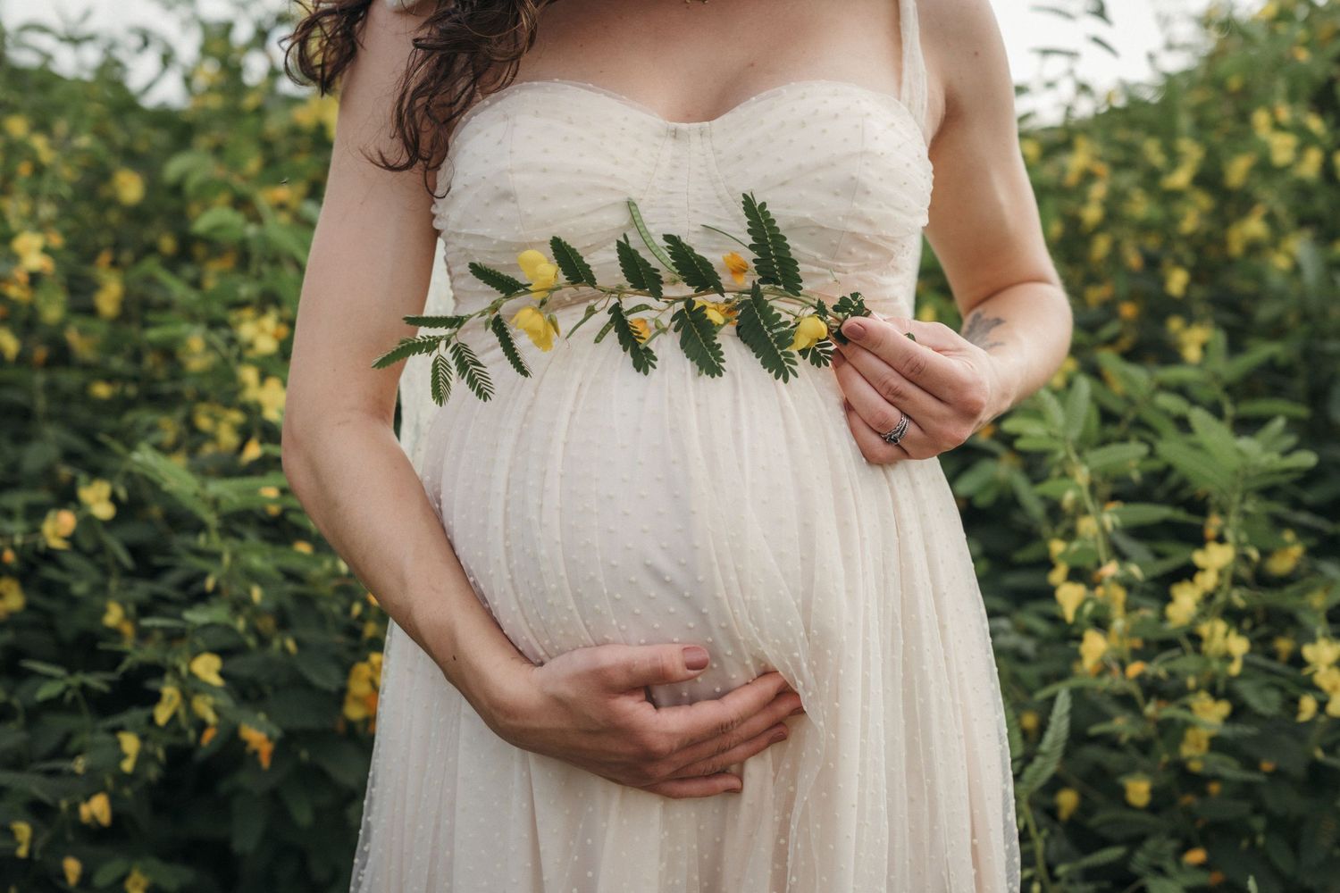 Pregnant woman in white dress holding yellow wildflowers against her baby bump in a garden at Seven Islands State Birding Park.