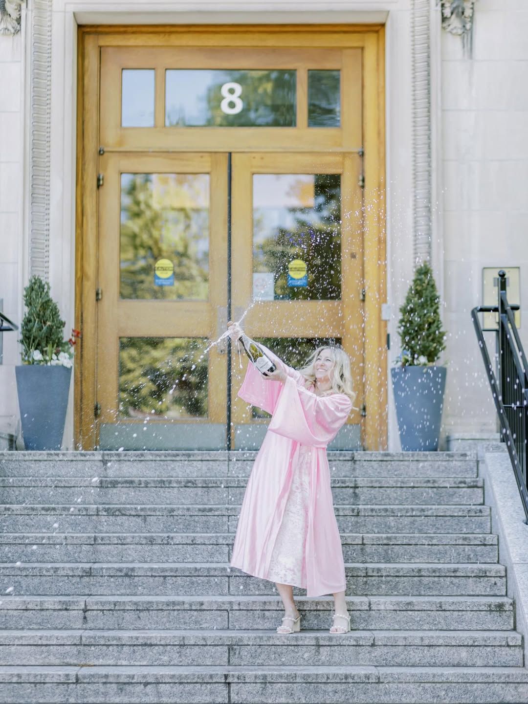 Graduate in pink gown pops champagne bottle on stone steps outside building number 8 in joyful celebration.