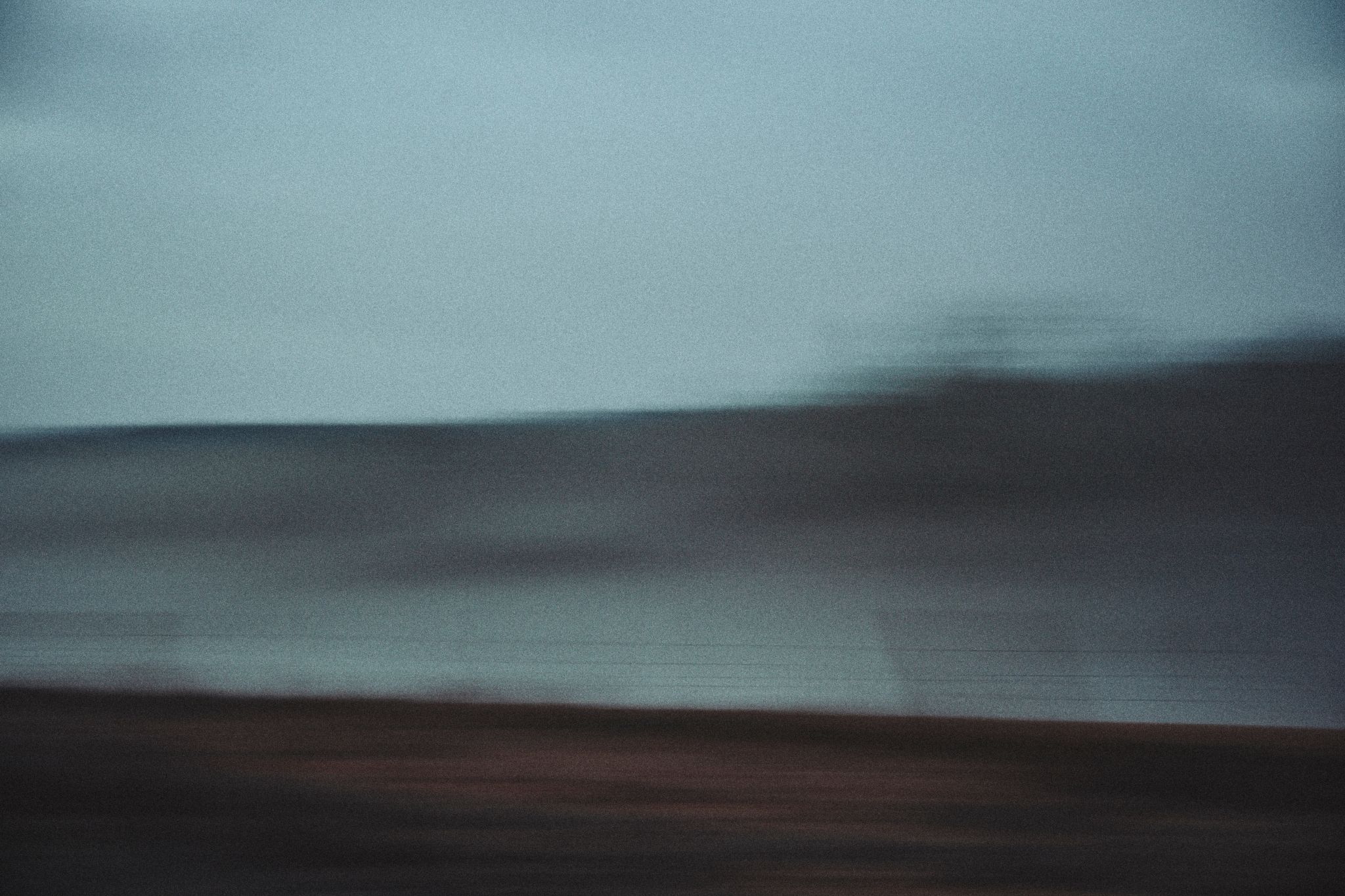 Abstract blurred landscape with moody blue-gray tones, motion blur effect creating a dreamlike coastal scene.