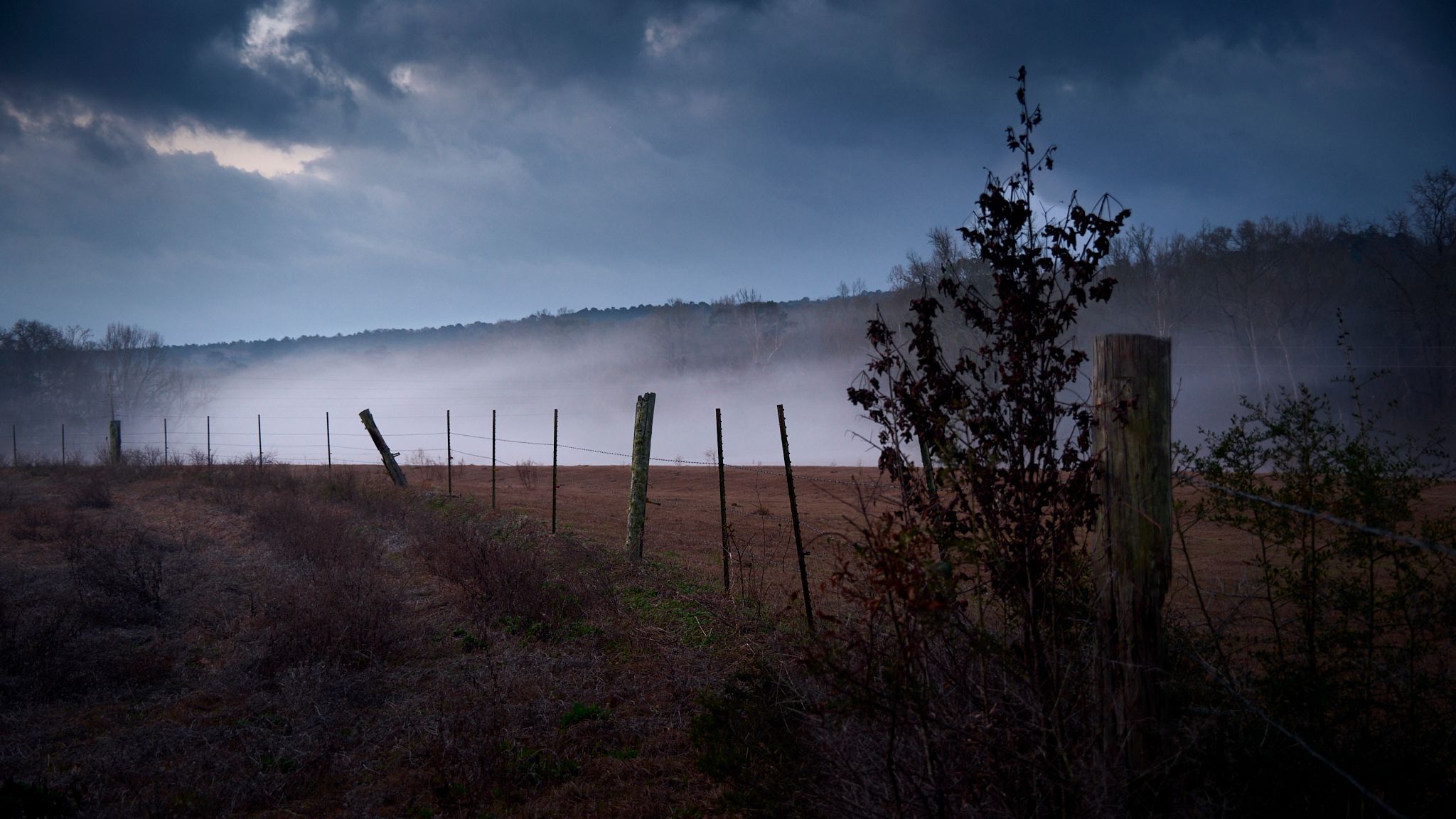 Misty rural landscape with wooden fence posts, barbed wire, and foggy hills under dramatic cloudy sky.