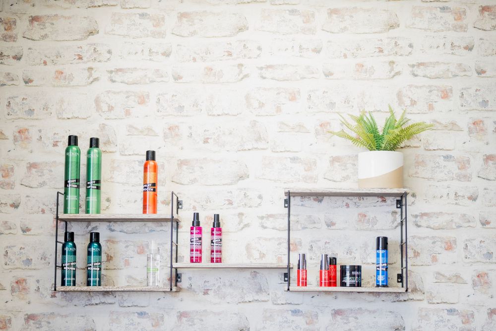 Hair care product bottles displayed on metal shelves against a white brick wall with a small plant.