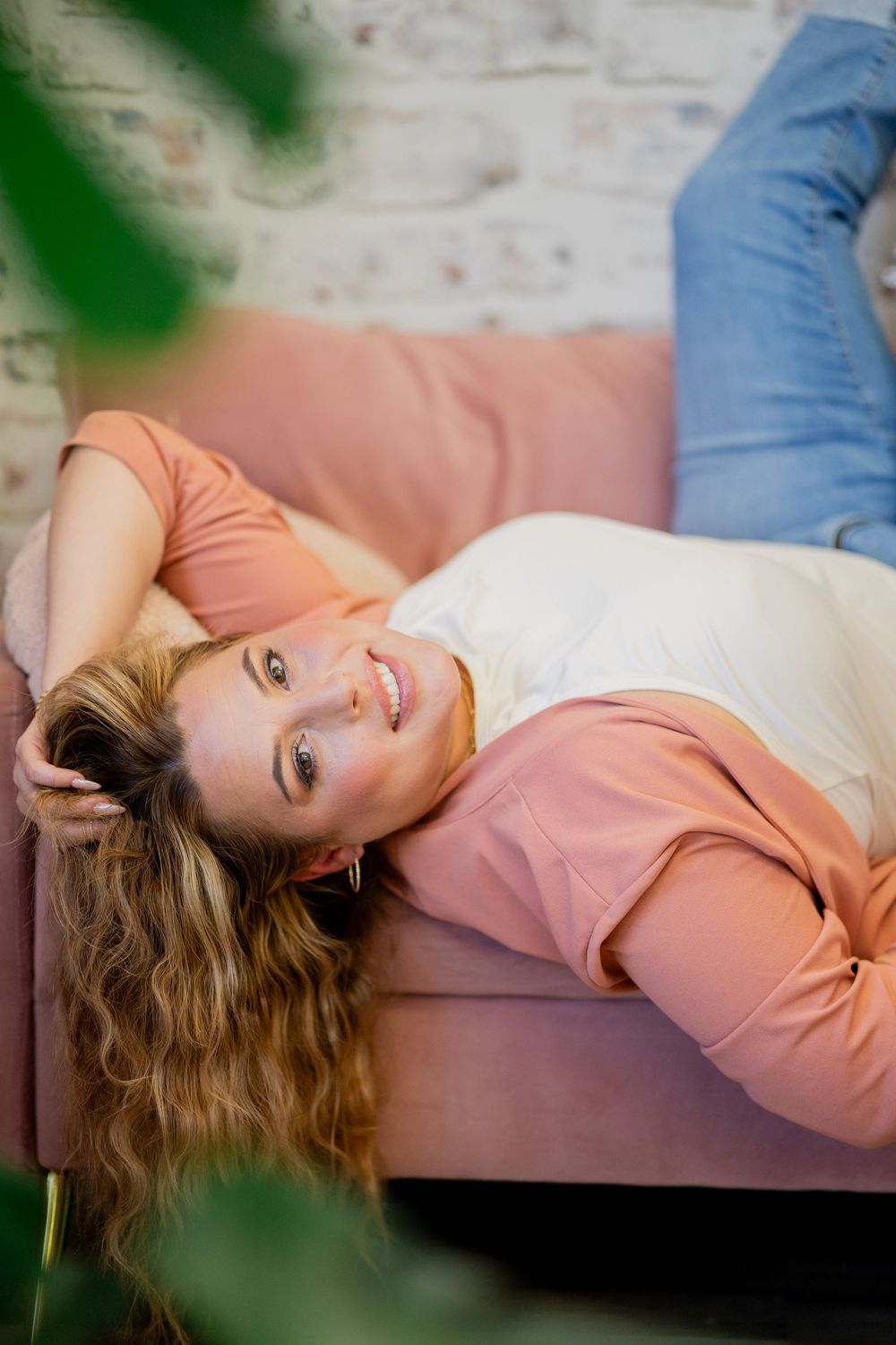 Smiling blonde woman relaxing on a pink velvet sofa viewed through green leaves.