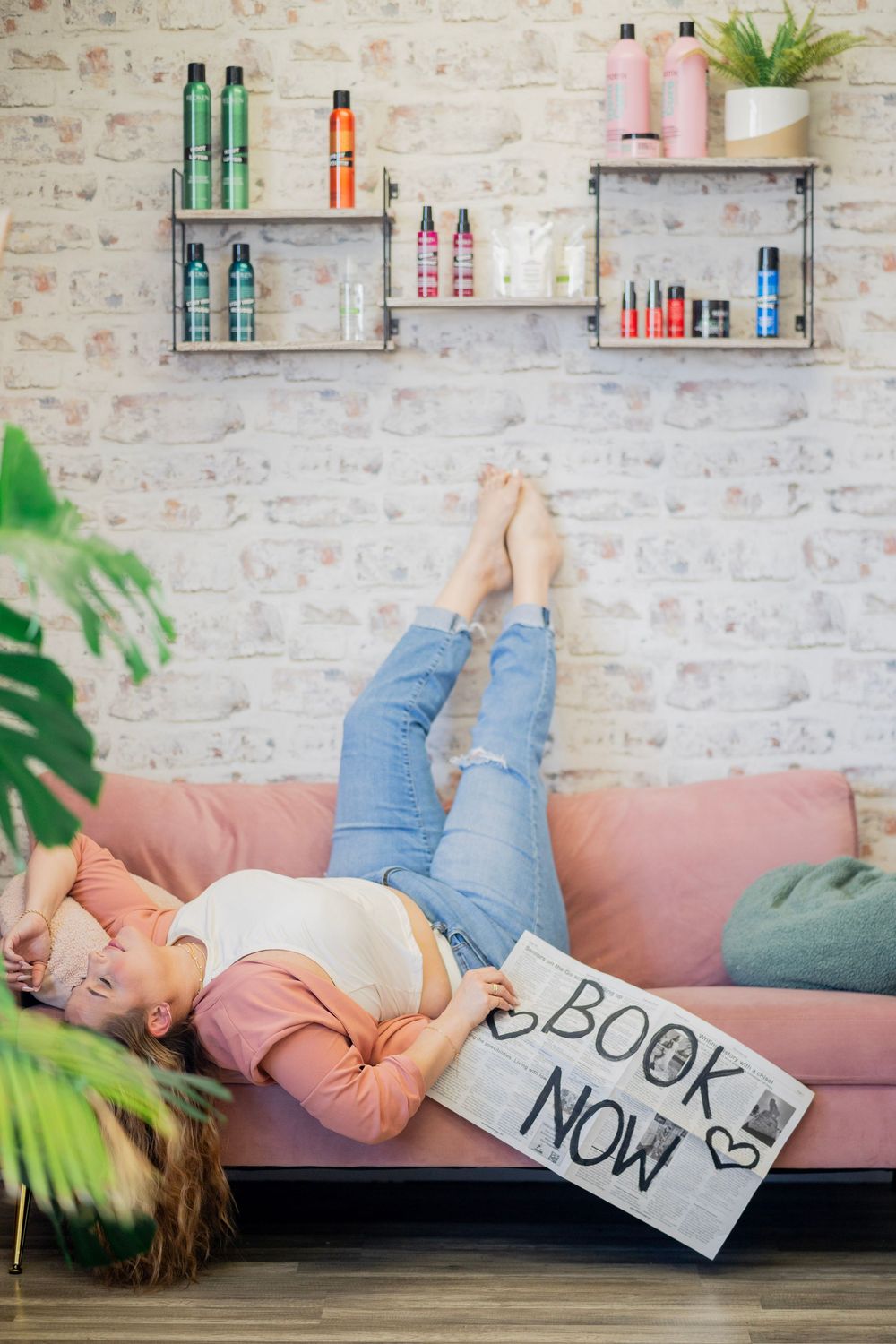 Woman lying on pink sofa holding Book Now sign with hair product shelves behind her.