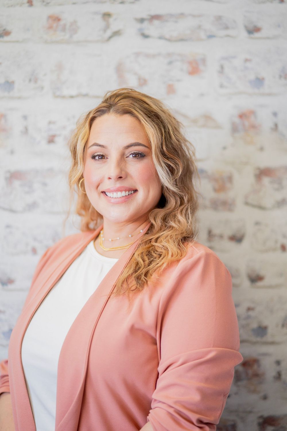 Smiling blonde woman in pink blazer posing against a white brick wall.