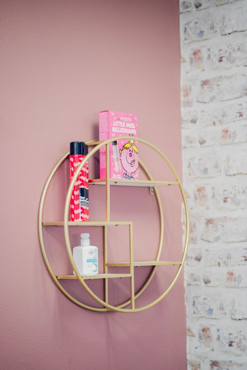 Gold circular wall shelf with pink beauty products and a Little Miss Billionaire book on a pink wall.