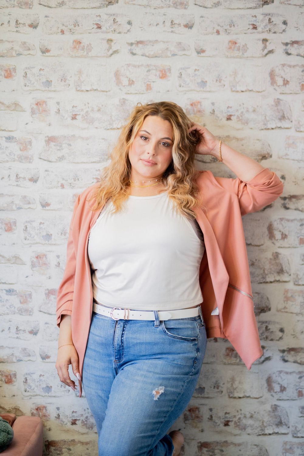 Confident blonde woman in pink blazer and jeans leaning against a white brick wall.