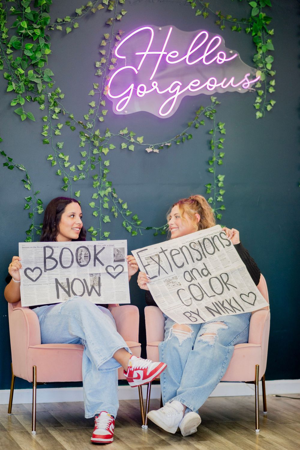 Two women smiling at each other holding Book Now and Extensions and Color by Nikki signs.