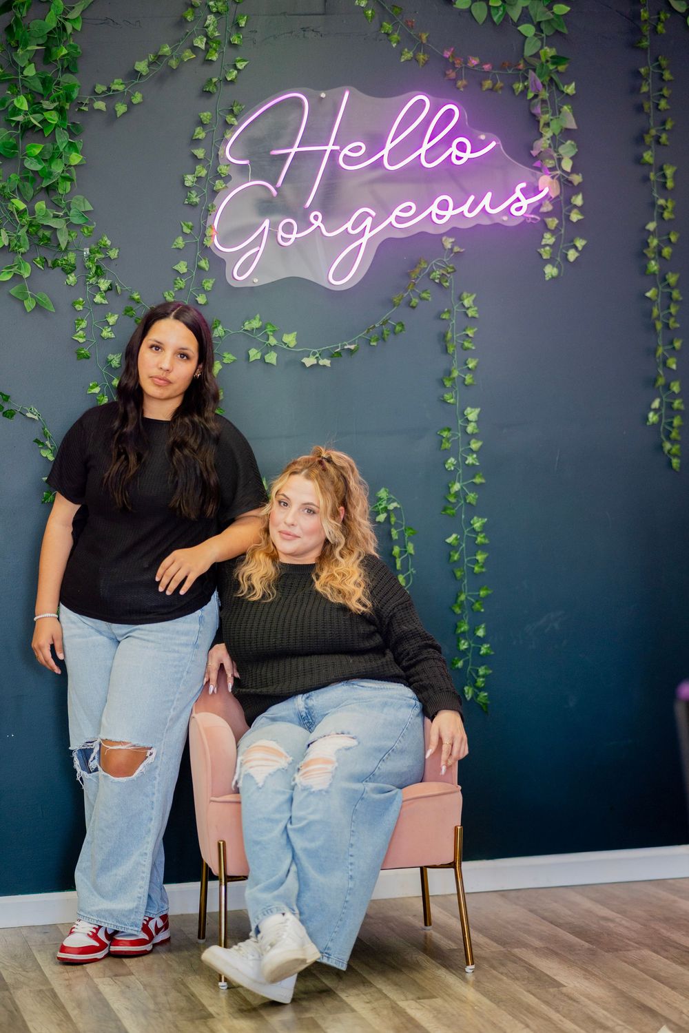 Two women pose in front of a neon Hello Gorgeous sign with ivy wall in a beauty salon.