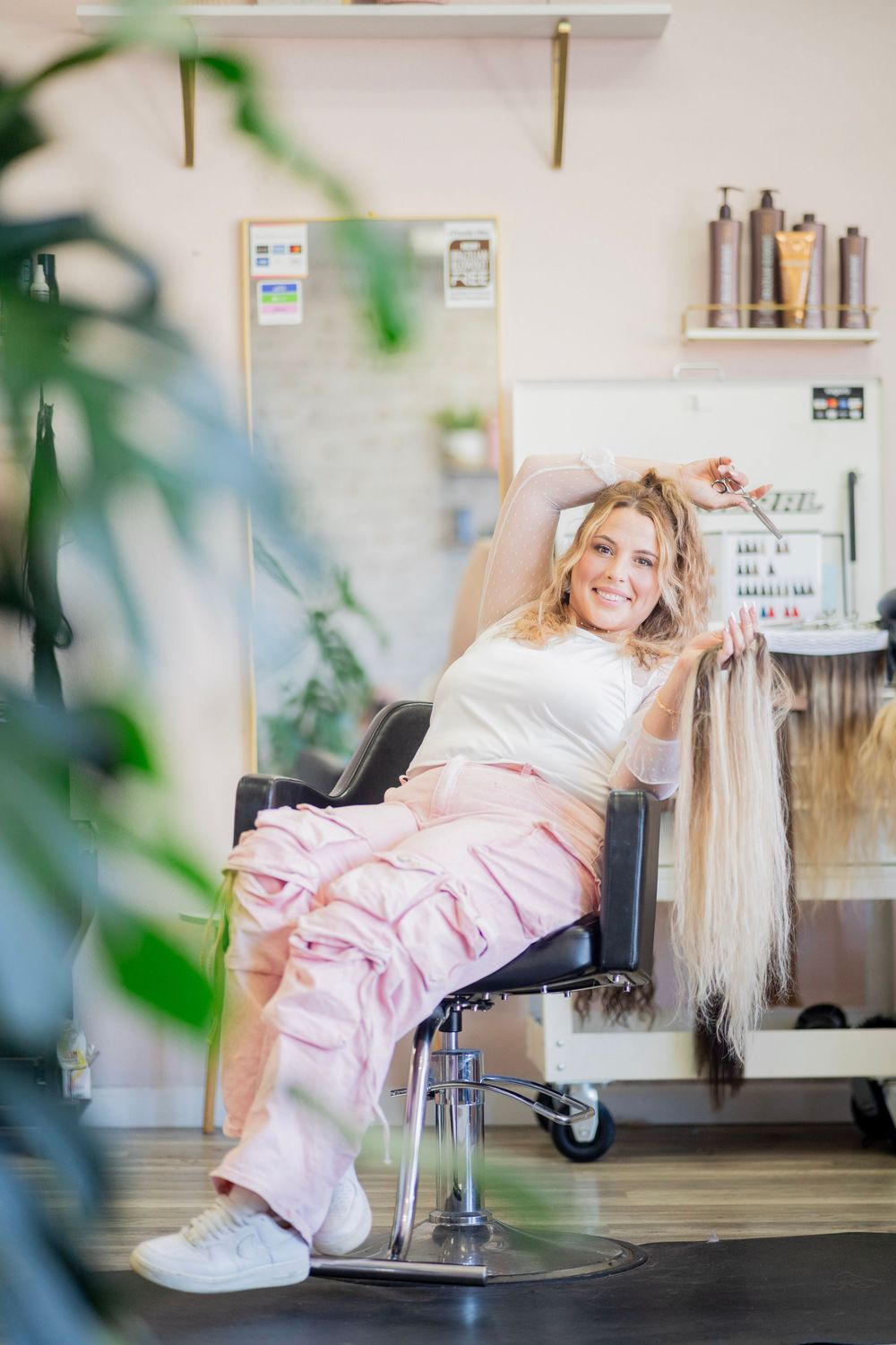 Happy blonde hairstylist relaxes in salon chair holding long hair extensions while smiling.