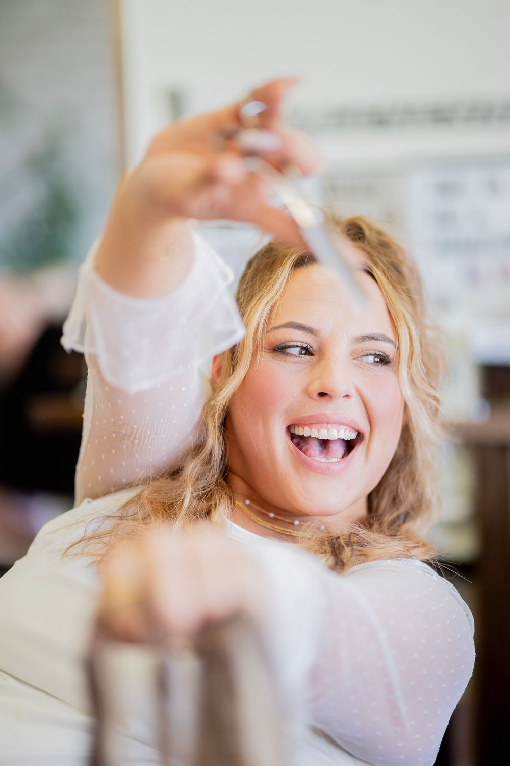 Laughing blonde woman holds scissors up in a bright hair salon environment.