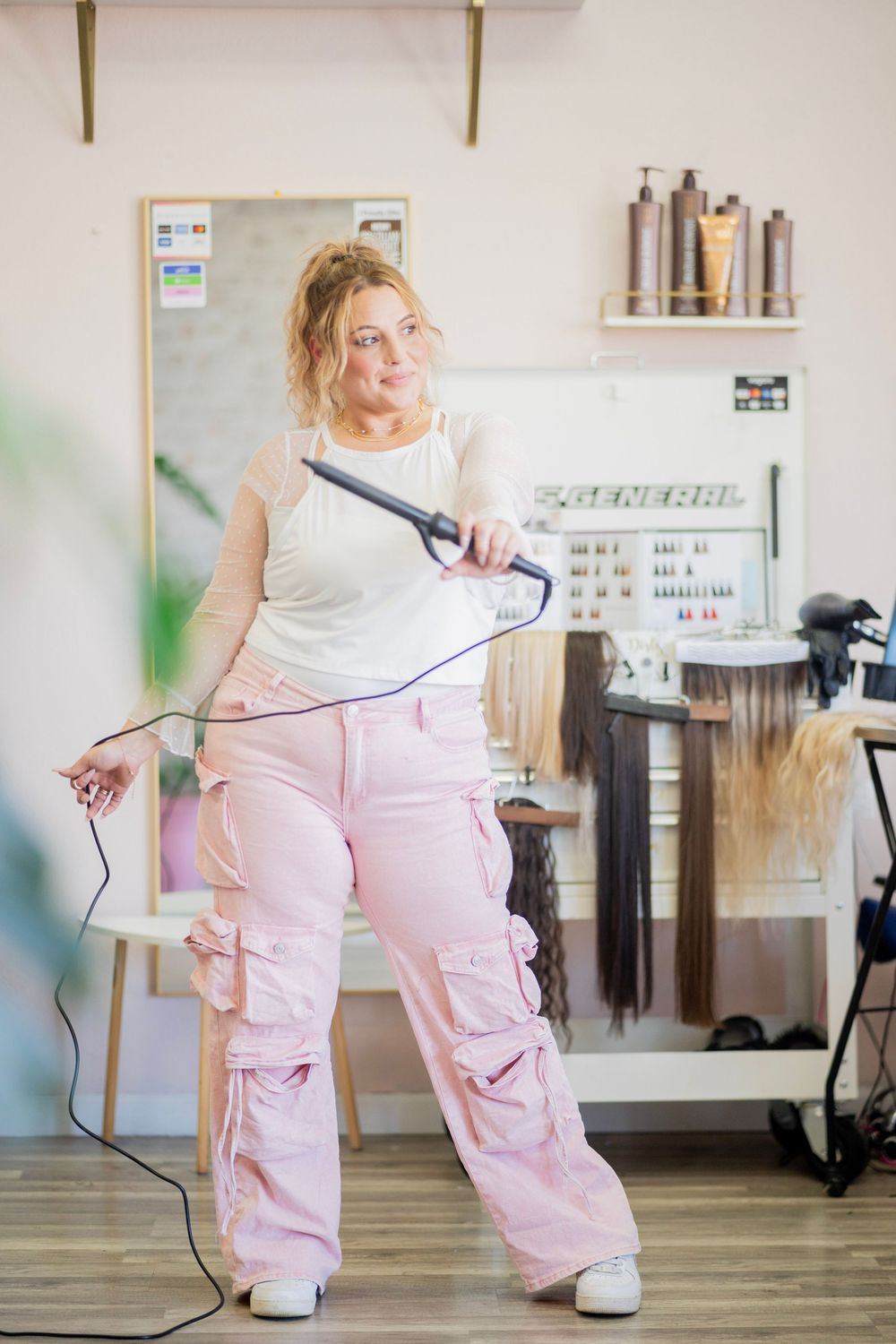 Blonde hairstylist poses with a curling iron in pink cargo pants in a hair salon.