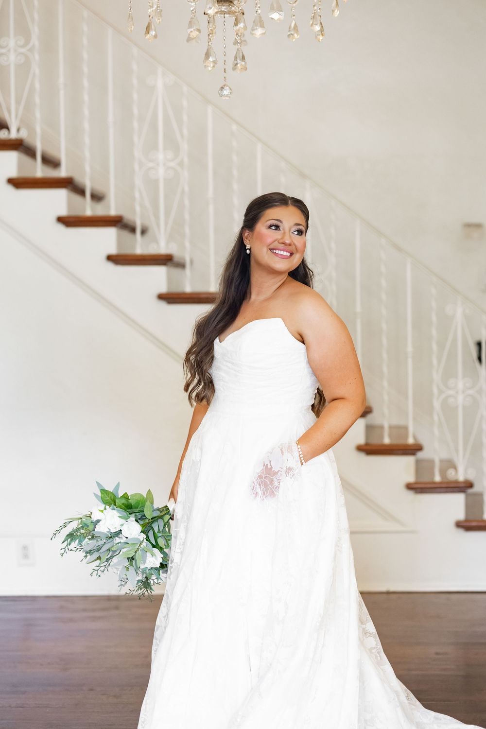 Radiant bride in strapless lace gown poses with white bouquet near elegant staircase with chandelier.