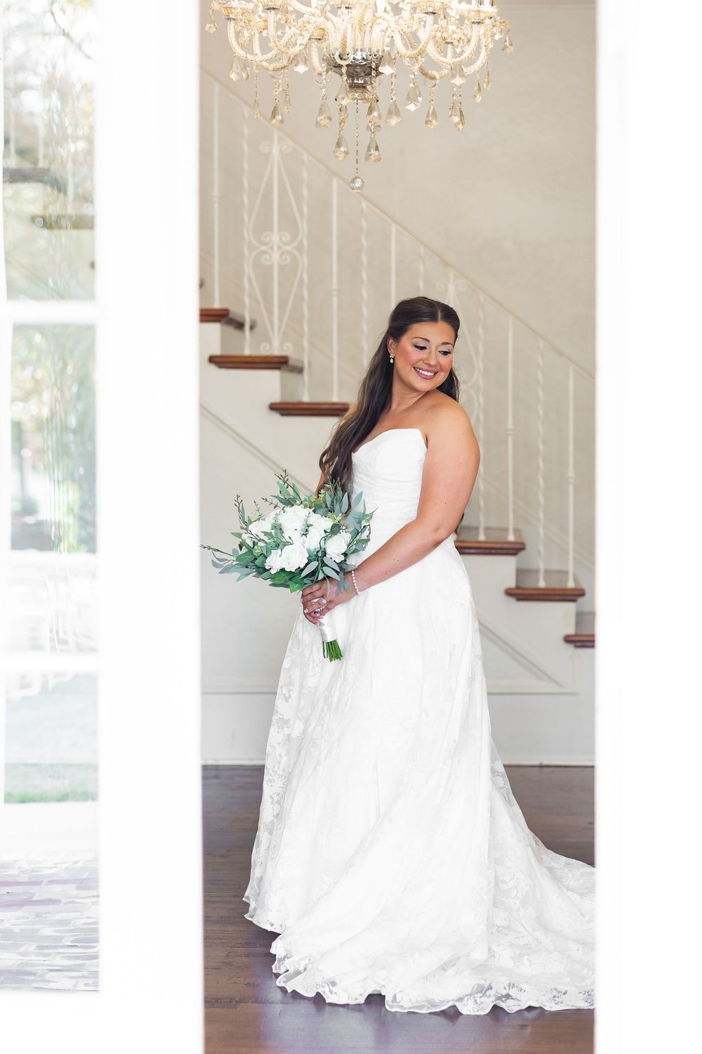 Beautiful bride in strapless lace wedding dress holding white floral bouquet beneath a crystal chandelier.