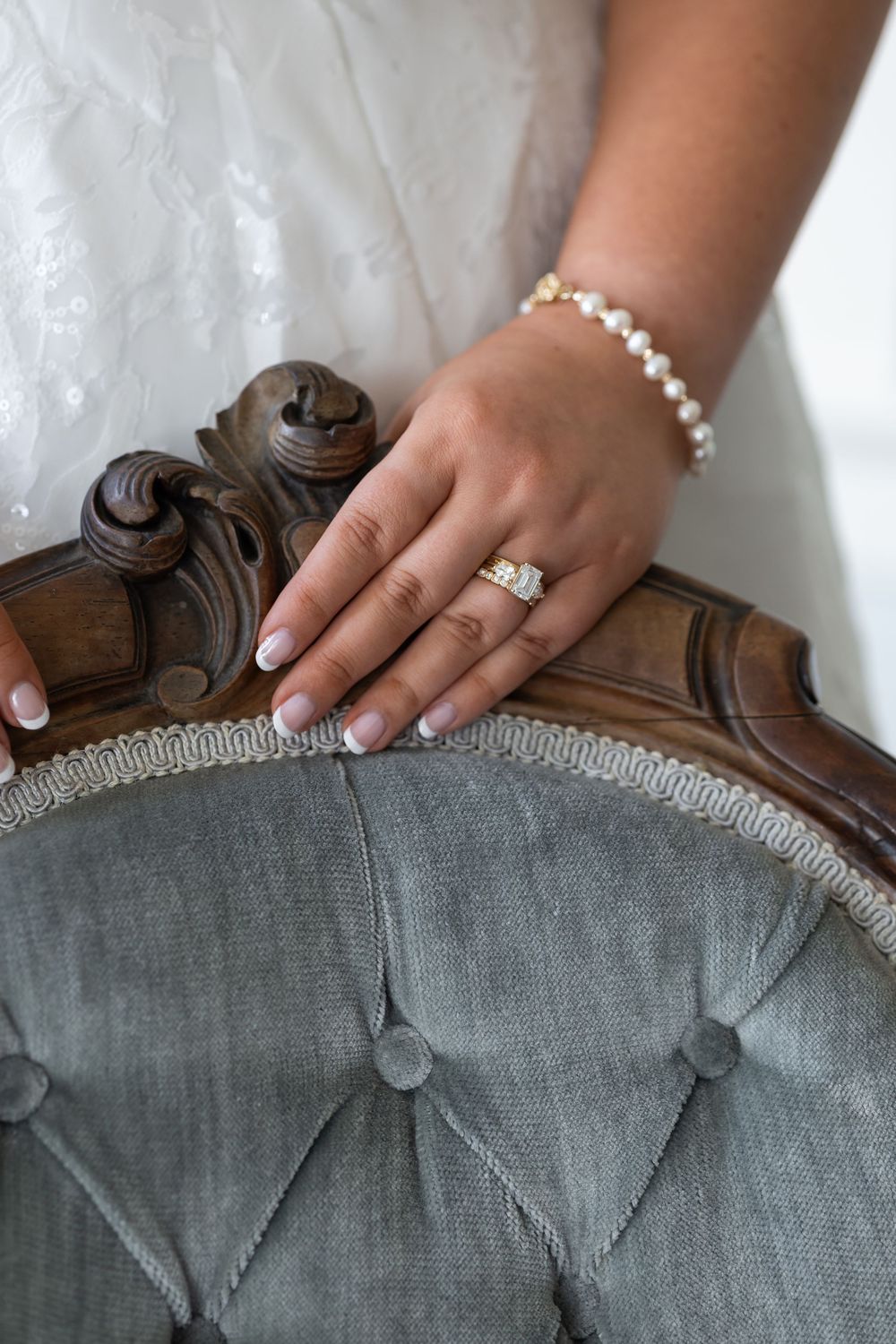 Bride's hand with diamond engagement ring and pearl bracelet resting on vintage tufted gray velvet chair.